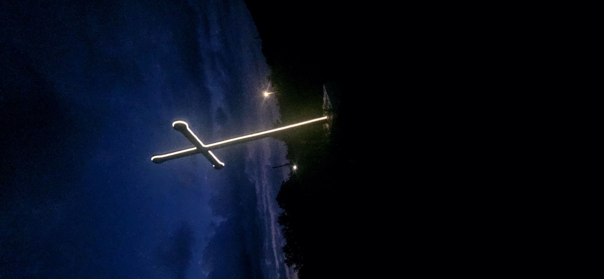 The image depicts a large, illuminated cross monument standing prominently against a dark blue, cloudy twilight or nighttime sky. The cross itself is outlined with bright, warm white or yellowish light, contrasting sharply with the dim environment. It appears to be constructed of a dark material, with the illumination tracing its edges. The cross is slightly tilted towards the right.
The upper portion of the frame is dominated by the dark blue sky, filled with heavy, dark clouds that show subtle variations in tone, indicating lingering ambient light or light from distant sources.
The midground and foreground are largely plunged into darkness, showing silhouetted terrain and dense vegetation, likely trees and bushes, on both the left and right sides of the frame. Two additional artificial light sources are visible in the background: one prominent, bright yellow streetlamp or light fixture is located in the mid-right, appearing further back than the cross, and a smaller, less intense light is visible within the dark vegetation on the left.
No individuals or discernible actions are visible in the scene. The image captures an outdoor setting, likely a hillside or elevated area, under low-light conditions. This illuminated cross is located in Gutsal, Bulgaria.