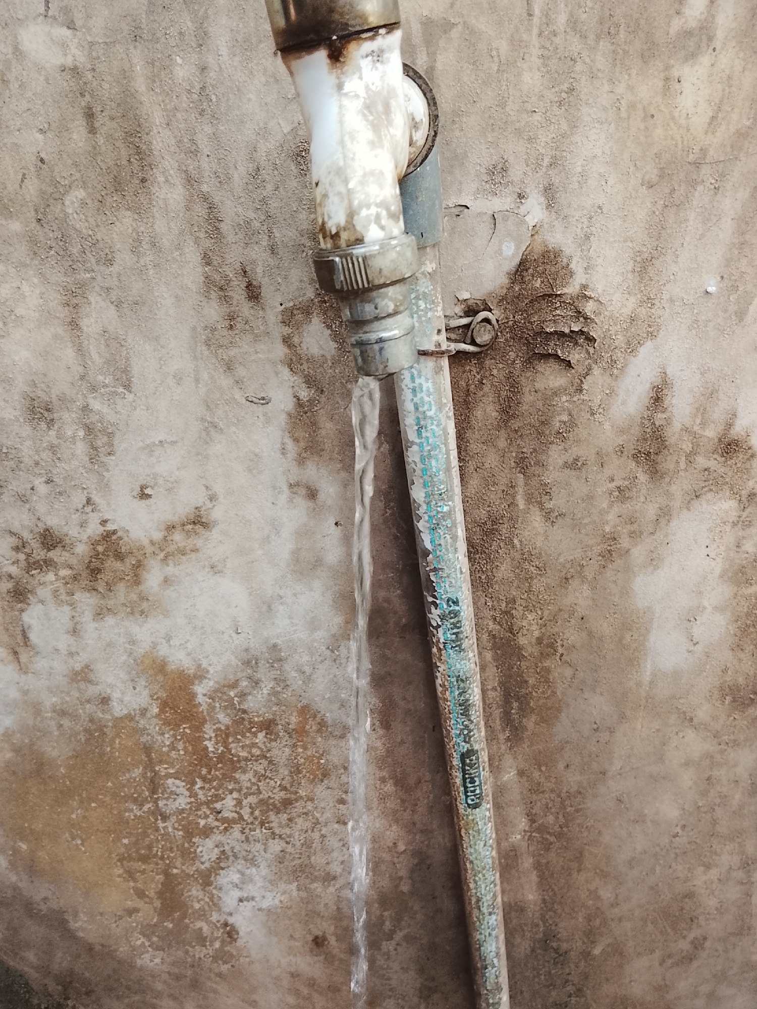 An outdoor scene shows a rusty pipe dripping water against a weathered, stained concrete wall. The pipe, possibly made of plastic and metal, is discolored with age and rust. A thin stream of water flows downwards from the pipe. The concrete wall displays patches of dirt, mold, and peeling paint, indicating a humid or neglected environment. The overall impression is one of decay and disrepair. The image is a close-up, focused on the pipe and the immediate wall surface, with no indication of the broader surroundings or specific location within Sukasejati, Indonesia.