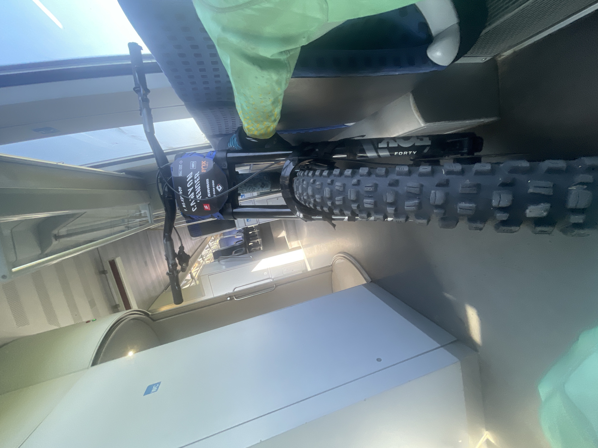 The image shows a front view of a mountain bike inside a train carriage. The bicycle is predominantly featured in the foreground, with its front tire, handlebars, and part of the frame clearly visible. A person wearing a bright green jacket is partially visible on the right side of the frame, their arm extended towards the bike. The background reveals the interior of the train, with seats, windows showing daylight, and a visible sign indicating a toilet ("WC") on the left. The scene appears to be set in Merichleri, Bulgaria, within the confines of a moving train.