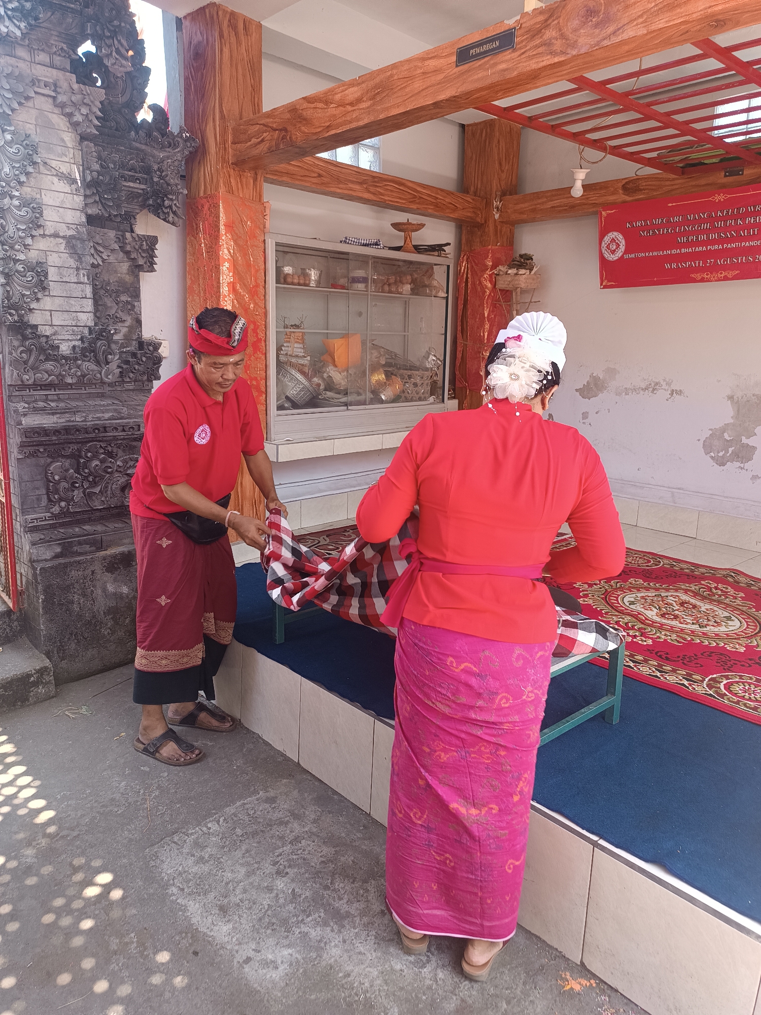 Two people are in the process of laying out a red and white checkered cloth on a blue carpeted platform. The person on the left, a man wearing a red polo shirt and a traditional red and black sarong, is holding one edge of the cloth. He has a black fanny pack around his waist and a red headband with a patterned design. The person on the right, dressed in a vibrant red long-sleeved top and a bright pink patterned sarong, is assisting with the cloth. She has a white headdress adorned with a floral ornament. The setting appears to be outdoors, possibly a temple or cultural space, with ornate stone carvings on the left and wooden beams overhead. A red banner with text is visible on the wall in the background, and a glass-fronted cabinet containing various items is also present. The ground in the foreground is concrete, with dappled light creating circular patterns. The scene is likely in Kerambitan, Indonesia, given the cultural attire and architectural style.
