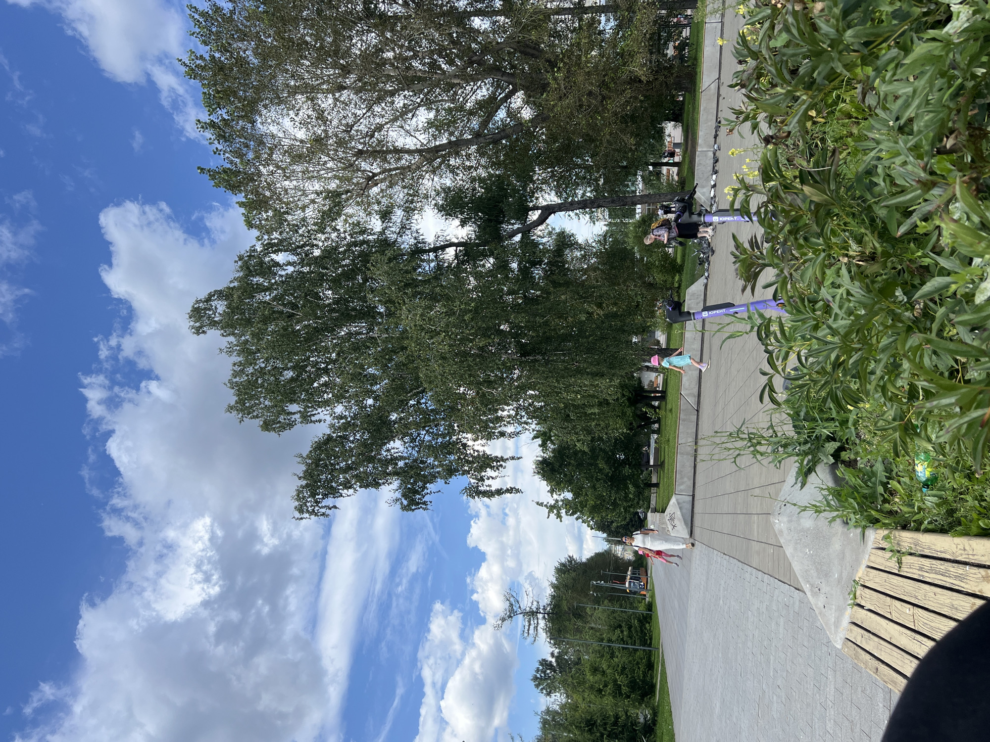 The image depicts a sunny day in Yekaterinburg, Russia, likely in a park or public square. A paved walkway stretches into the distance, flanked by lush green trees and a cloudy blue sky. In the foreground, a wooden bench sits beside a bed of green plants. Several people are visible on the walkway; a woman in white walks away from the camera, a child in a pink hat runs ahead, and another child in a blue dress follows. Further down the path, a woman with a backpack stands near several parked electric scooters. A tram can be seen in the background, and a flock of pigeons congregates on the ground near the scooters. The overall scene conveys a sense of leisure and outdoor activity in a city park setting.