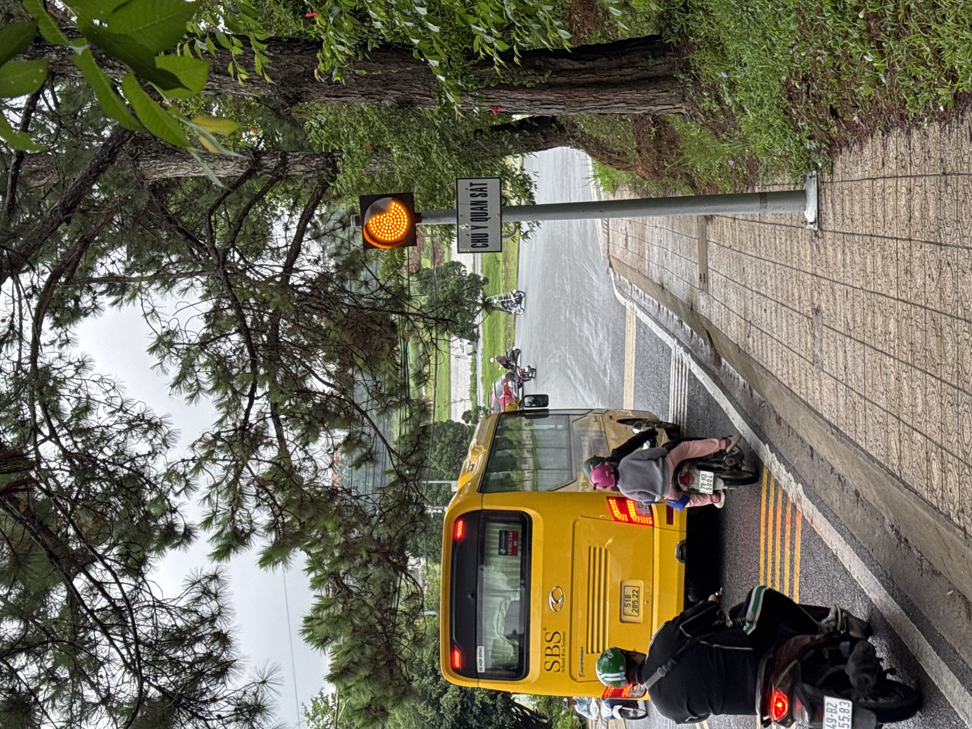 The image depicts a road scene in Phường Xuân Hương - Đà Lạt, Vietnam, under an overcast sky. The road surface appears damp, suggesting recent rainfall.
In the foreground, a bright yellow bus, which has "SBS School Bus System" written on its side, is stopped on the right side of the road. Its brake lights are illuminated. Several motorbikes are also stopped around the bus. One motorbike, with a rider wearing a pink helmet, is positioned to the left rear of the bus, while another, with a rider in a green helmet, is directly behind the bus.
To the left of the bus and closer to the foreground, a traffic pole stands with a yellow/amber blinking light at its top, indicating a cautionary signal. Below the light, a white rectangular sign with black lettering is affixed. The right side of the road features a textured sidewalk, which then gives way to a grassy embankment. Large trees with dense green foliage, likely pine trees, canopy over the scene, with branches extending into the frame.
The visible text on the sign reads "CHÚ Ý QUAN SÁT," which translates to "ATTENTION OBSERVE" or "PAY ATTENTION." On the yellow bus, the text "SBS School Bus System" is prominent, along with "VŨNG TÀU" and the license plate "51B 285.22." A motorbike in the foreground displays the license plate "49-B2 55.83," and another motorbike near the bus has the license plate "79-K3 500.4." The word "HỢP ĐỒNG" (Contract) is also visible on the bus. The scene captures a moment of halted traffic, typical of an intersection or a specific crossing point.