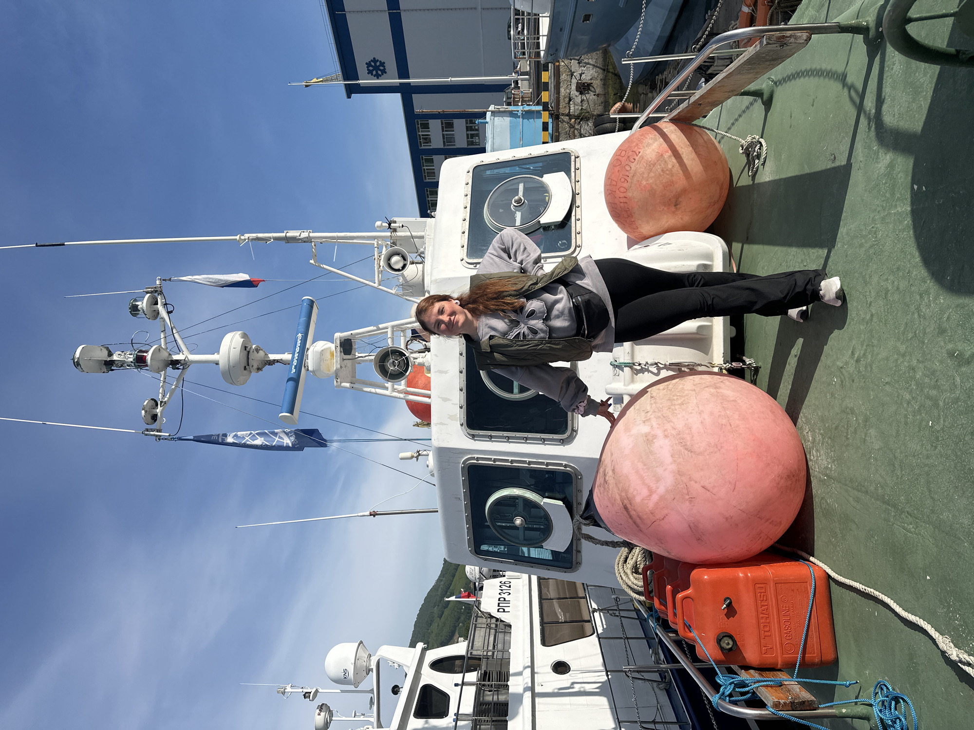 A young woman with auburn hair is posing on the deck of a white boat, with a larger boat to her left. She is wearing a gray sweatshirt, a dark green vest, black pants, and gray sneakers. A black fanny pack is worn around her waist. She has her left hand on her hip and her right hand by her side. The deck is painted green and has large pink buoys and orange fuel cans. The boat has large windows and is equipped with navigation equipment and flags. In the background, another boat and a building with a blue roof are visible. The image appears to have been taken in a harbor in Petropavlovsk-Kamchatsky, Russia, as indicated by the presence of Russian flags and the overall maritime setting consistent with the city's location on the Pacific coast. The sky is blue with some clouds.