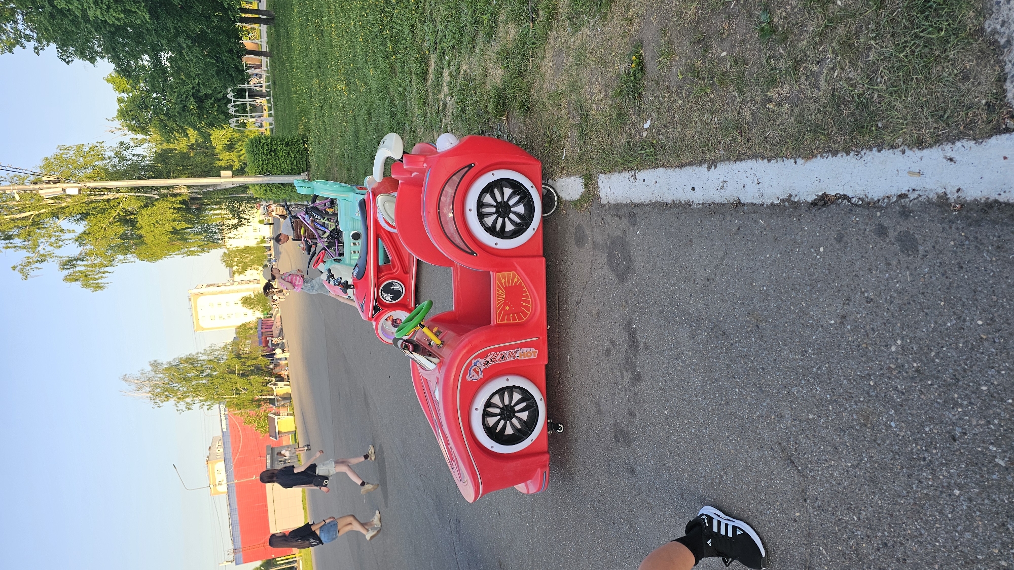 The image shows an outdoor scene on an asphalt surface, likely a street or sidewalk. A red, toy car-like structure, potentially an amusement ride, is prominently featured on the right side of the frame. A white stripe marks the edge between the asphalt and a grassy area. The grassy area has a few weeds.
Several individuals can be seen walking on the street in the distance. The nearest visible person's foot and shoe, black with white stripes, are visible in the foreground.
In the background, buildings and trees are visible under a clear sky.