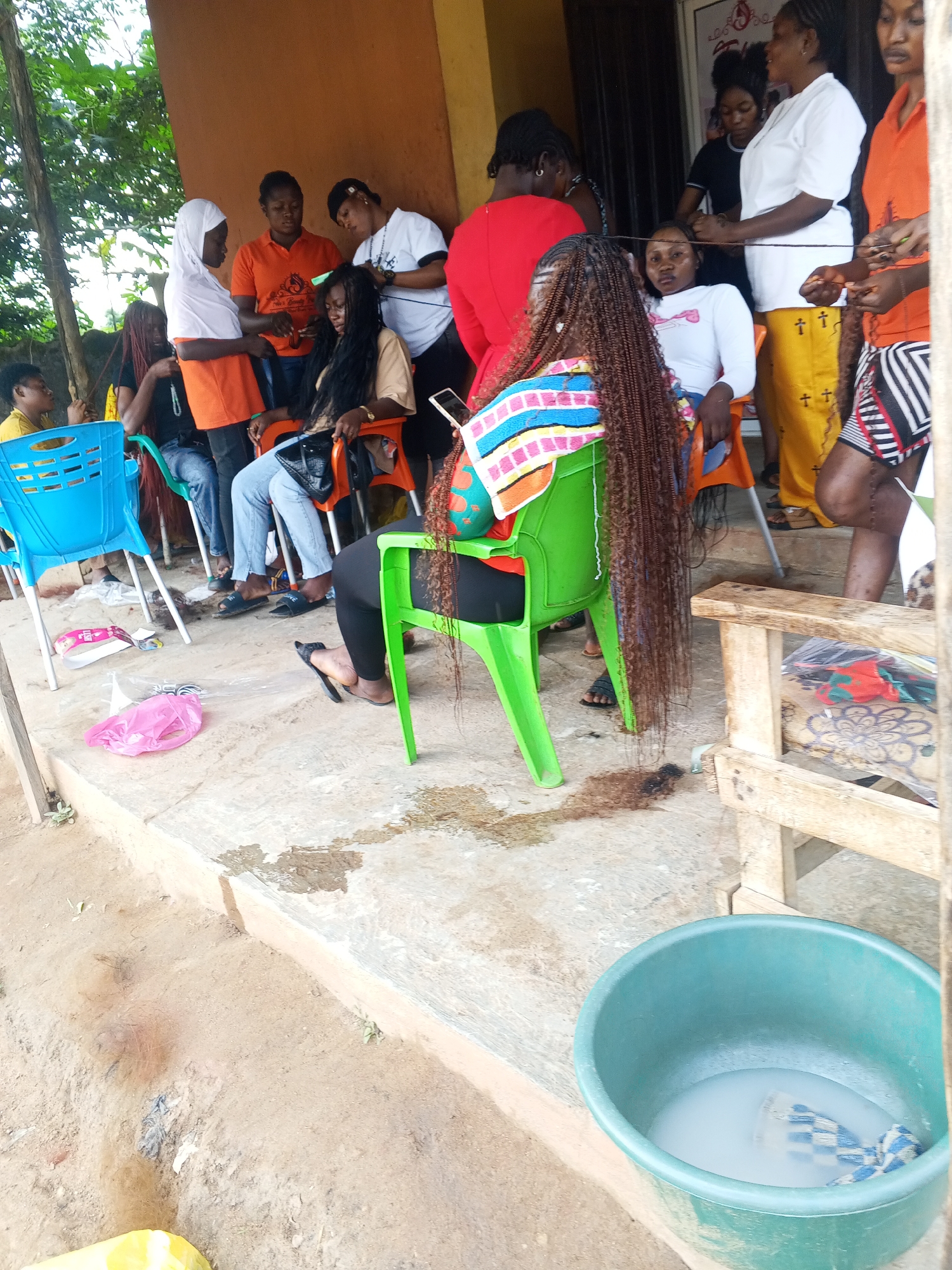 The image depicts an outdoor scene in Kabba, Nigeria, where a group of women are gathered, seemingly for hair braiding services. In the foreground, a woman with long, braided brown hair is seated in a green plastic chair, her back to the camera. Her hair extends down her back and onto the floor. To her left, a blue plastic chair is empty.
A short distance away, other women are visible. One woman, wearing a white headscarf and an orange top, stands and appears to be attending to the braids of another woman seated in an orange chair. Several other women are also seated or standing in the background, engaged in conversation or activities related to hair styling. Some are wearing casual attire, including t-shirts and patterned skirts.
The environment appears to be a somewhat informal outdoor setting, possibly in front of a building with an orange-colored wall. The ground is a mixture of concrete and dirt, with some puddles of water and scattered debris, including a pink plastic bag. A large green tub filled with soapy water is positioned on the ground in the lower right portion of the frame, suggesting preparation for washing hair or materials. The overall atmosphere suggests a communal and practical setting for a hair salon.