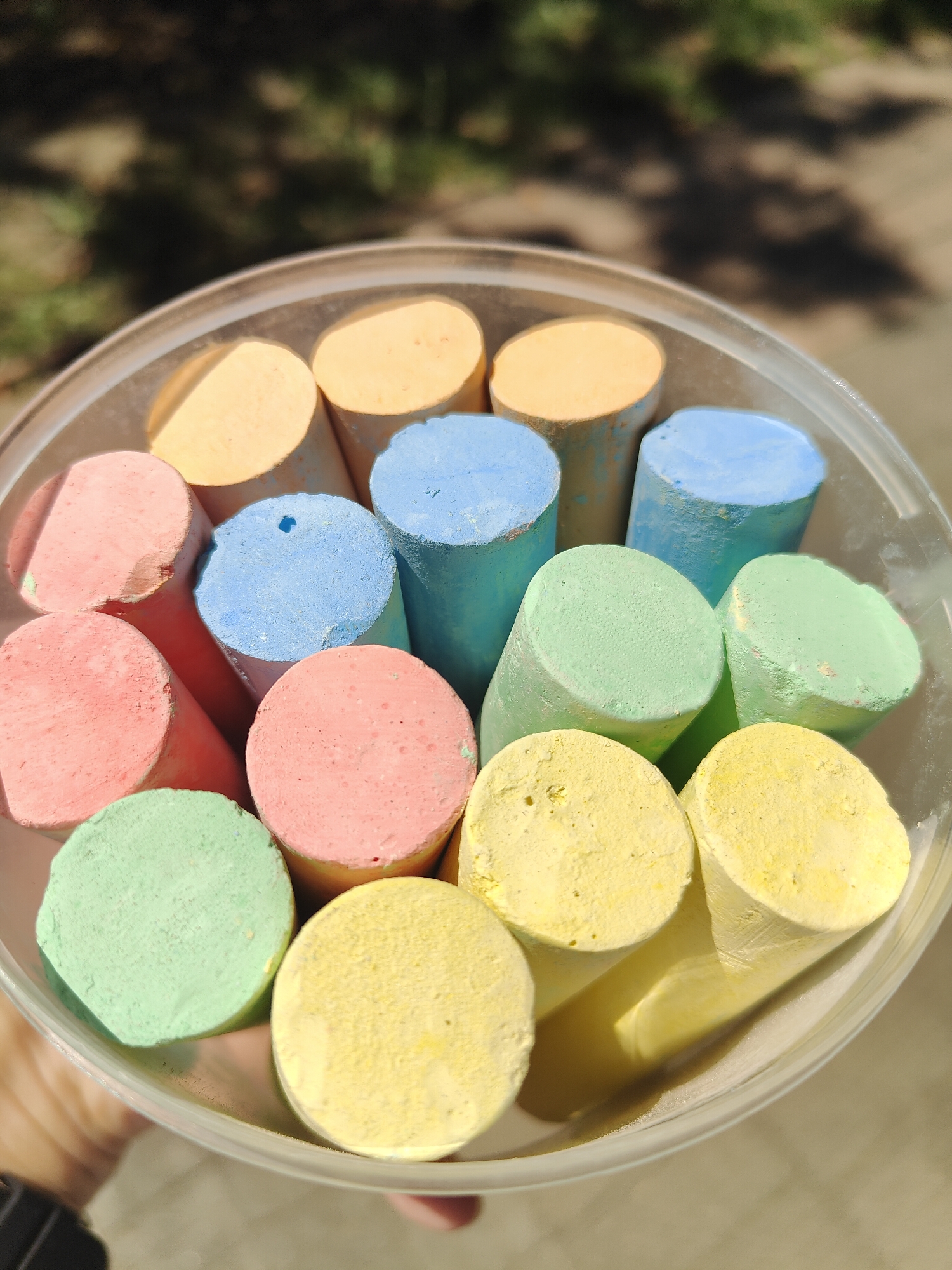 A close-up, overhead view shows a clear plastic container filled with chunky, cylindrical sticks of sidewalk chalk in various pastel colors, including yellow, orange, pink, light blue, and light green. The chalk sticks are arranged in a somewhat haphazard manner, with some standing upright and others tilted. The container is held in a person's hand, with a dark watch visible on their wrist. The background is blurred and appears to be outdoors, with dappled sunlight filtering through trees, suggesting a park or residential area. The lighting indicates it is daytime. No specific landmarks or architectural features are visible to pinpoint a location within the city of Chornomorsk, Ukraine.