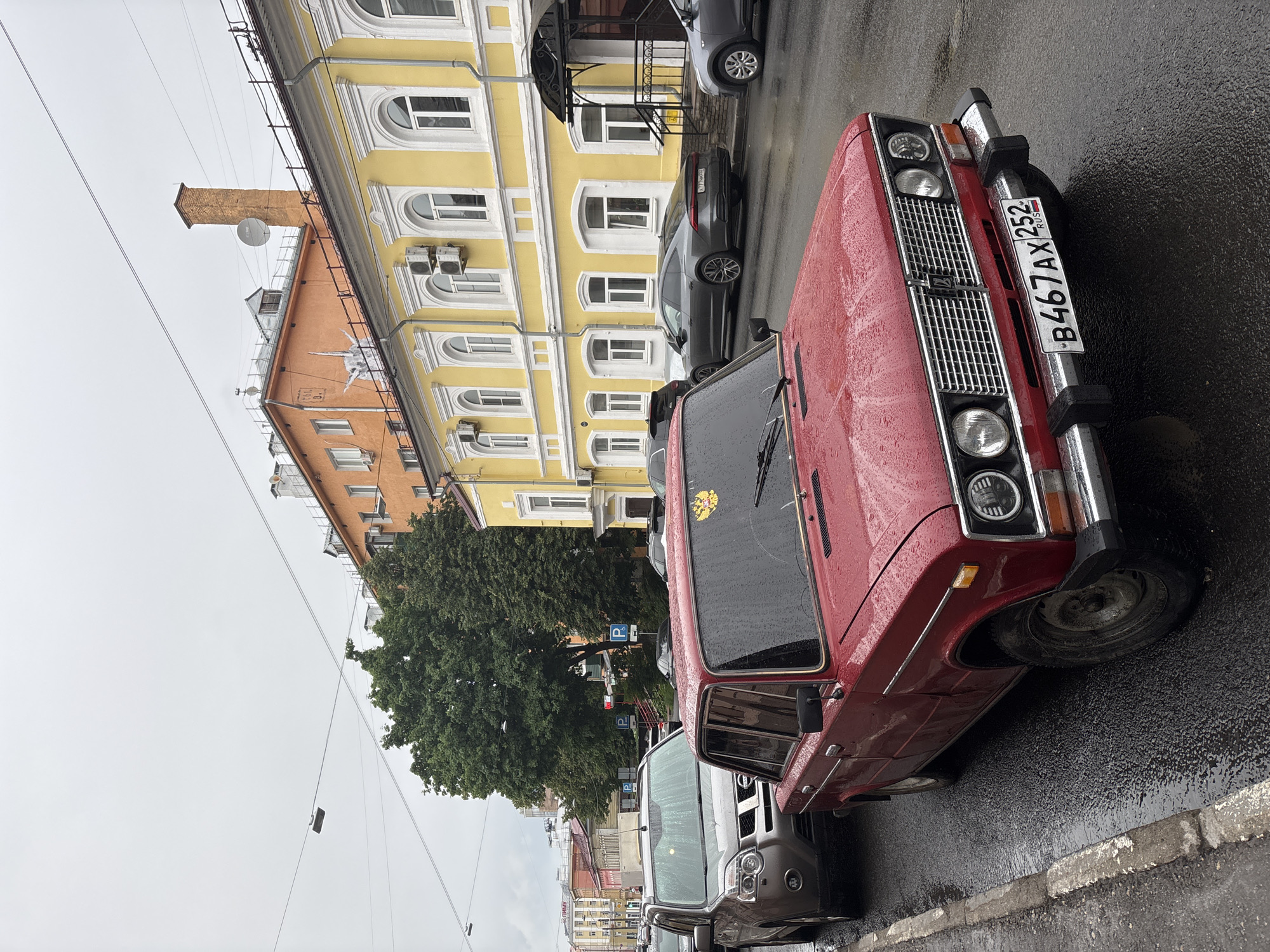 In Nizhny Novgorod, Russia, on a rainy day, a red vintage car is parked on a wet asphalt street. The car is a Soviet-era sedan, possibly a Lada, with a license plate that reads "B467AX 252". A silver SUV is parked behind it. In the background, a yellow building with arched windows and white trim stands prominently. Further back, orange and lighter-colored buildings are visible, along with lush green trees and utility poles. The sky is overcast, contributing to the somber mood of the scene. There are no people visible in the image.