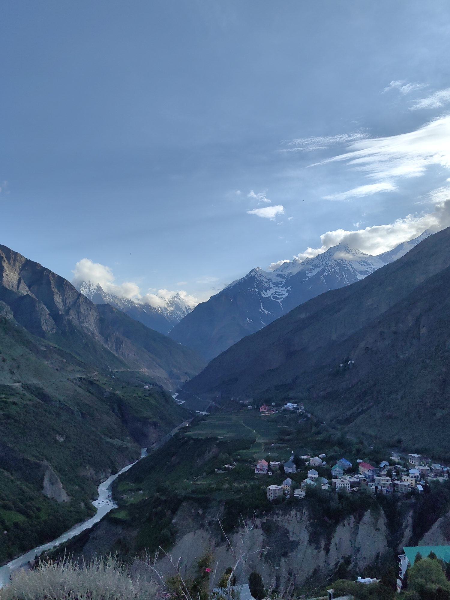 A wide, high-angle shot captures a valley in Keylong, India, under a partially cloudy blue sky. The valley is framed by steep, dark green mountains that show patches of sunlight and shadow. A wide, light blue river snakes through the valley floor, bordered by lighter green terraced fields. Nestled on the side of the valley, a cluster of buildings forms a small village with colorful roofs. In the distance, snow-capped mountain peaks rise above the clouds. The foreground shows rough terrain with some dry vegetation.