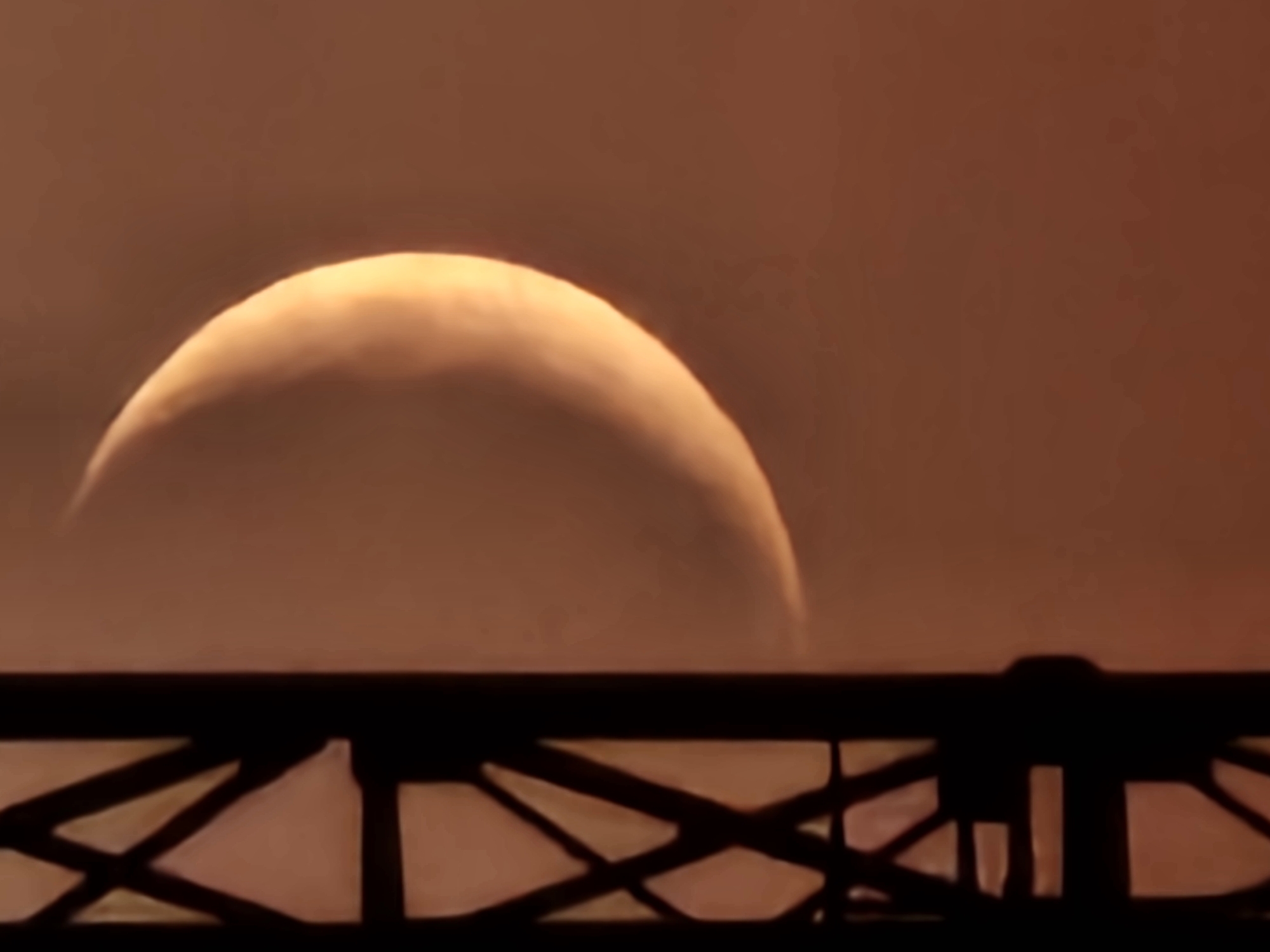 A crescent moon is visible in the sky, partially obscured by a dark, lattice-like structure on the left side of the frame. The moon is illuminated from the right, creating a bright, golden crescent shape against a muted, hazy, reddish-brown sky. The structure on the left appears to be made of dark metal or wood and is a vertical element within the composition. The overall impression is of an astronomical event observed from an urban setting, likely in the city of Khimki, Russia. The specific location within Khimki is not discernible from the image itself, but the presence of the structure suggests it might be part of a building, a tower, or some other urban architectural feature. No people are visible in the image.