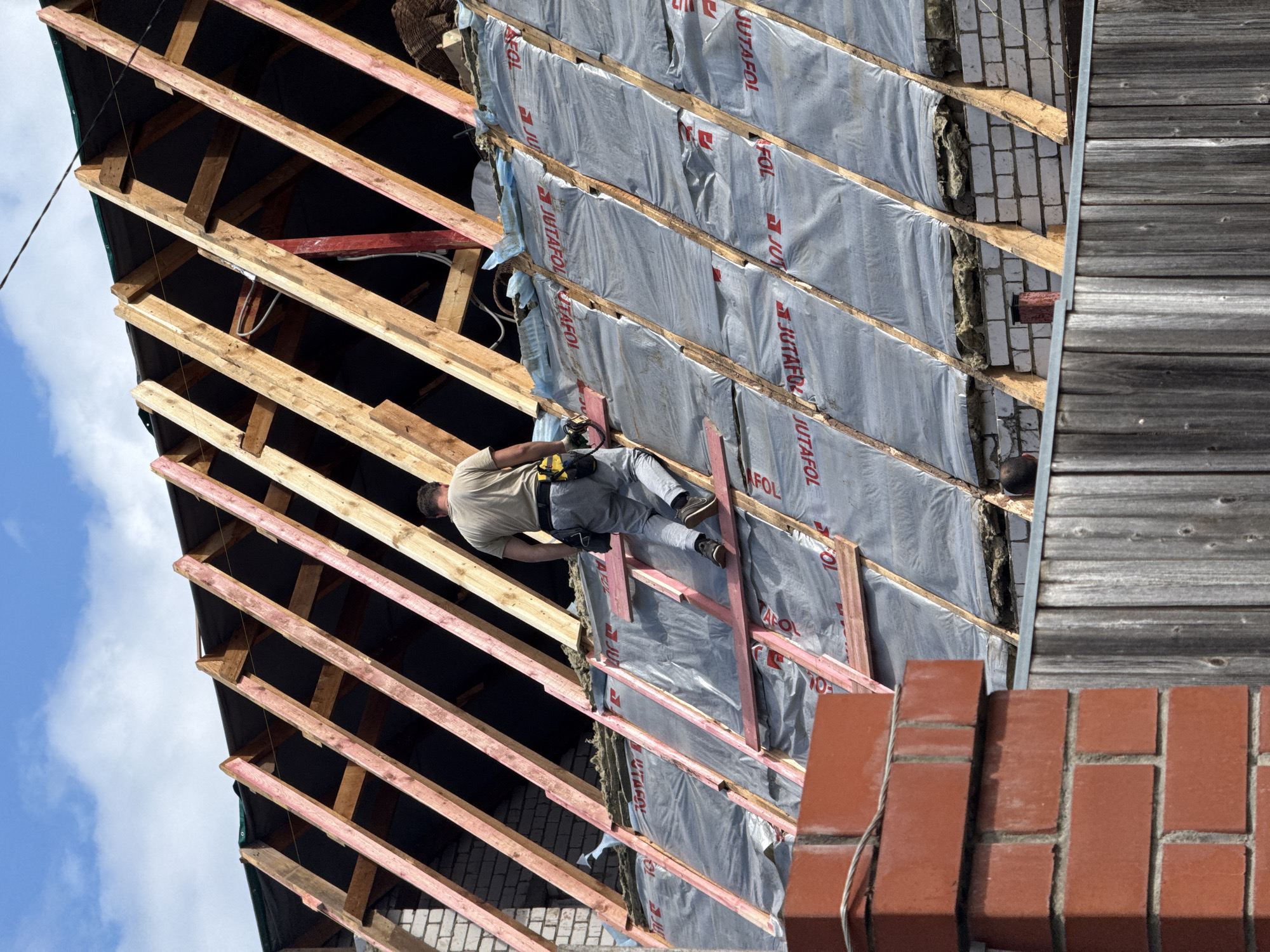 The image depicts a construction site, likely in Dmitrov, Russia, focusing on the exterior of a building undergoing roof work. A single construction worker, dressed in a light-colored t-shirt, gray pants, and a tool belt, is positioned on a slanted roof. The worker is in the process of ascending or maneuvering along the roof structure, which consists of wooden rafters and is partially covered with a silver, reflective vapor barrier. The roof's framing is visible, showing the underlying wooden beams. In the foreground, a red brick chimney or pillar and a weathered wooden fence are present, suggesting the property boundary. The background reveals a partly cloudy blue sky. The scene is characterized by the ongoing renovation or construction of the building's roof.