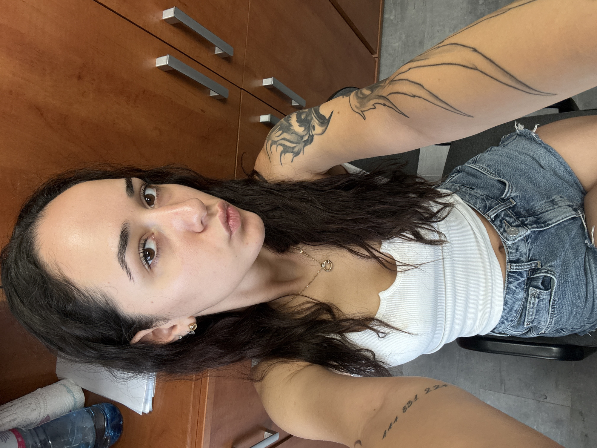 The image shows a woman with dark hair and light skin taking a selfie in what appears to be an office environment. She is wearing a white tank top and denim shorts. She has tattoos on both arms and is wearing earrings and a necklace. In the background, there are wooden cabinets with metal handles, and to the left, a bottle of water and some papers are visible on a desk. The floor is tiled. The scene is well-lit, likely with artificial lighting. The woman is looking at the camera with her lips puckered.