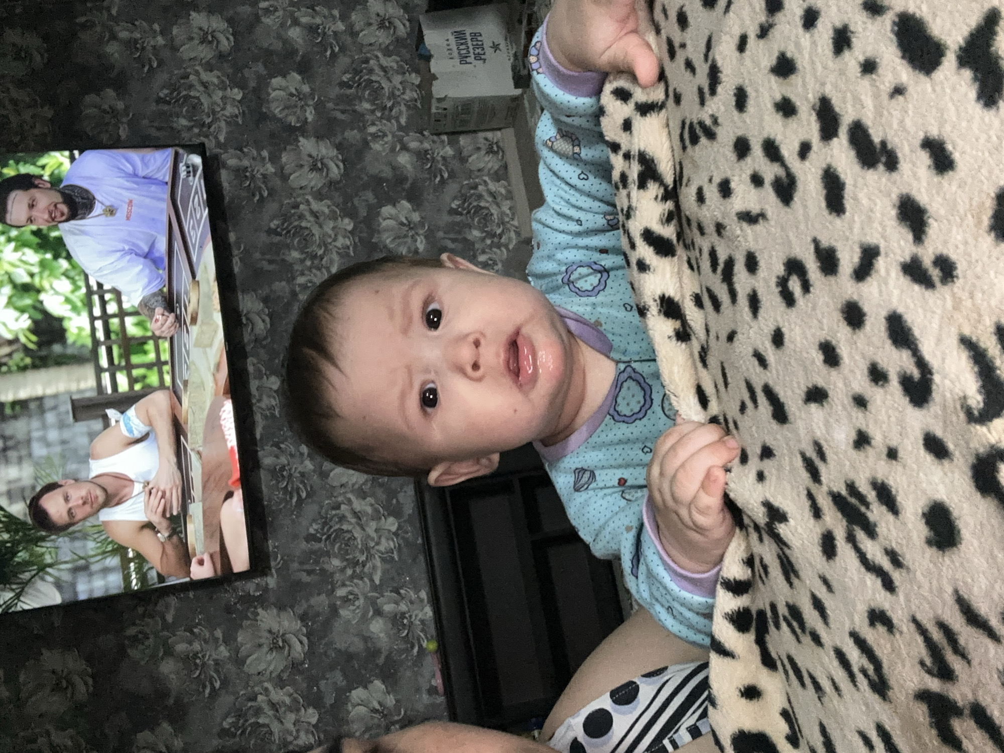 The image, oriented vertically, prominently features a baby's head and upper torso in the center. The baby, likely an infant or young toddler, has dark eyes and a slightly open mouth, looking directly at the camera with a neutral, curious expression. They are dressed in a light blue long-sleeved top adorned with small, light-colored patterns such as hearts and circles. The baby's left hand grasps a thick, cream-colored blanket or fabric with distinct black leopard spots, which fills much of the right side of the frame. Below the baby, parts of an adult's leg or thigh are visible, clad in striped and polka-dotted fabric, suggesting the baby is being held or is resting on an adult.
The background consists of dark floral wallpaper featuring intricate grey flower designs. To the left, a flat-screen television is displaying two different men. The upper man, with a beard and visible tattoos on his arms, is wearing a white t-shirt and a gold necklace, looking towards the left side of the screen. Below him, another man in a white tank top, also with tattoos, appears to be holding a guitar or similar stringed instrument. Both men on screen are set against an outdoor backdrop with lush green foliage.
Above the baby's head, resting on a dark surface against the wallpaper, is a cardboard box. Clearly visible Cyrillic text on the box reads "ВОДКА РУССКИЙ РЕЗЕРВ" (Vodka Russian Reserve), along with smaller, less legible text beneath it. The scene depicts an intimate indoor setting, likely a home, with the baby and an adult engaged in watching television. The time of day is not discernible from the indoor lighting.