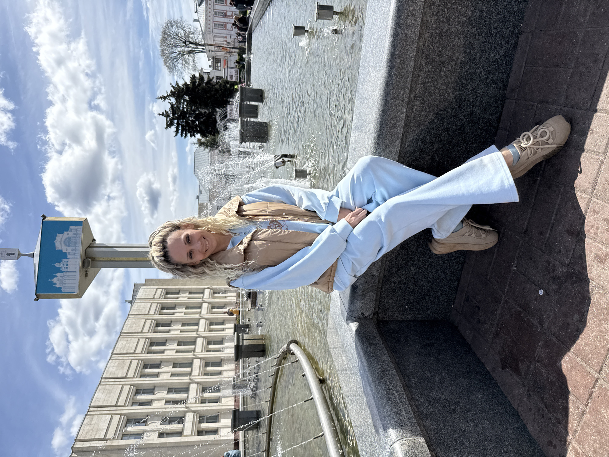 A woman is sitting on the edge of a fountain. She has blond hair and is wearing a light blue sweatsuit with a tan vest and tan sneakers. The fountain is located in a city, with a large building in the background and a signpost with a blue and white graphic to the left of the woman. The sky is blue with white clouds. The location is Unknown, Unknown.