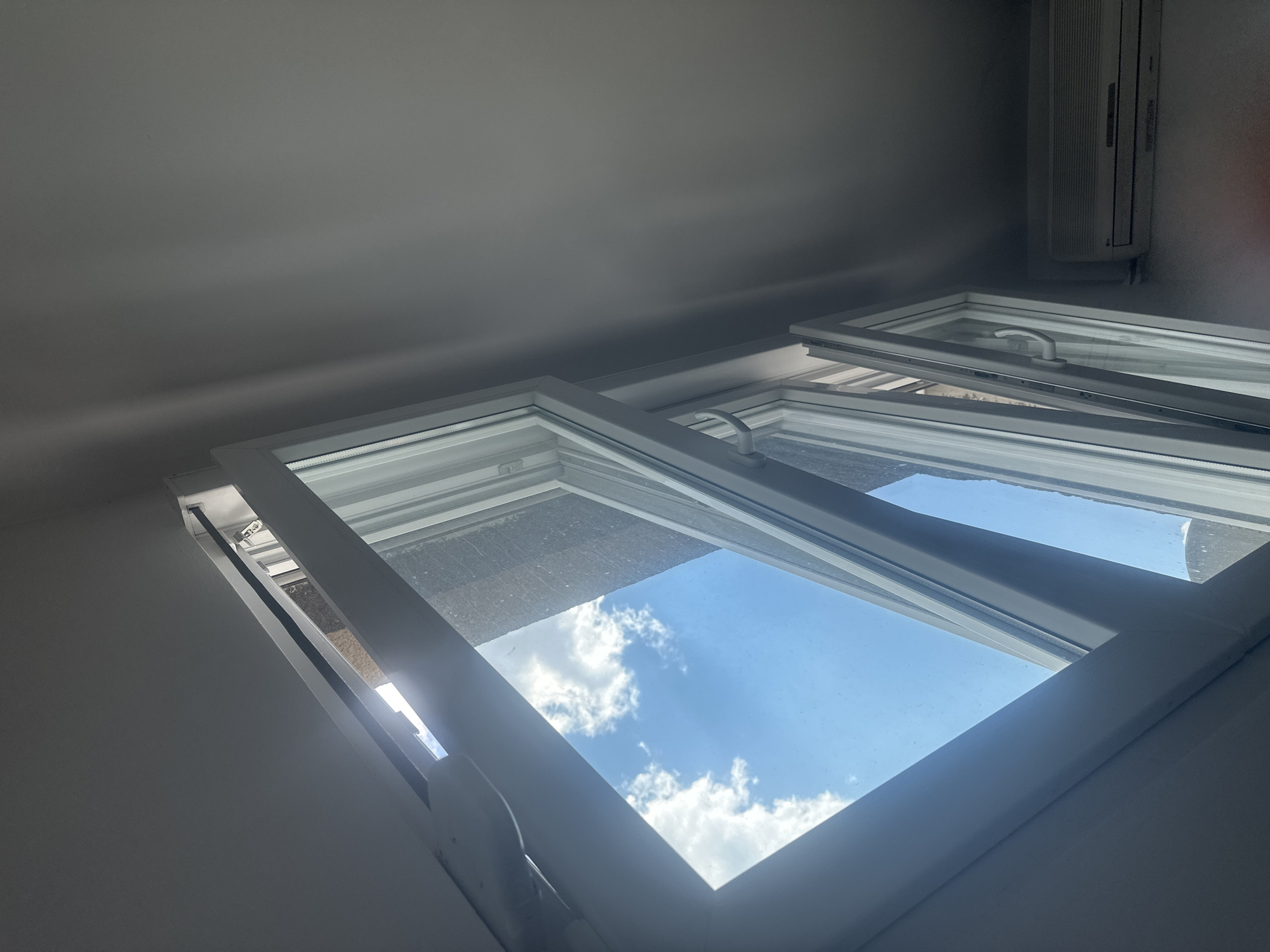 The image shows an interior view of a room looking up at an open window. The window is white and appears to be a tilt-and-turn style. The top portion of the window is tilted inwards, allowing a view of a bright blue sky with fluffy white clouds. The sunlight streams through the opening, casting shadows on the interior walls and ceiling. The walls are a neutral, light color and appear smooth. Mounted high on the wall above the window is a white air conditioning unit. The overall impression is that of a bright, sunny day, possibly late morning or early afternoon. The scene is serene and peaceful, with the open window suggesting ventilation and a connection to the outdoors. No people are visible in the image. The location is identified as Zvezdara Urban Municipality, Serbia.