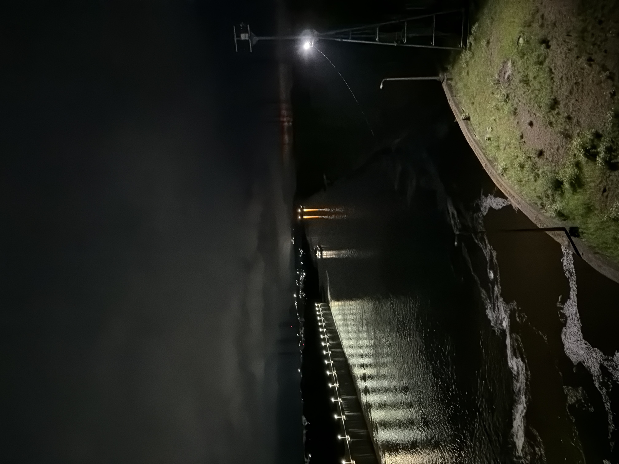 This image displays a detailed nighttime scene of a section of the Kama River in Chaikovsky, Russia, adjacent to a significant hydraulic engineering structure. On the left side, a long concrete dam or lock wall extends into the distance, illuminated by a series of evenly spaced streetlights that cast prominent, elongated reflections across the dark river surface. A motorized vessel, showing visible internal or deck lighting, navigates the waterway, creating its own distinct light reflections. Further downstream on the right side of the river, additional orange-red light reflections are visible. The immediate right bank features a tall, four-legged metallic tower, likely a navigational aid or monitoring station, equipped with a powerful spotlight directed downward onto the water. A smaller, conventional streetlight is also present on this bank. The foreground on the right consists of a curved, vegetated earthen embankment with a concrete retaining edge, bordering the river where patches of white foam and turbulent water are discernible. The upper portion of the image reveals a dark, cloudy night sky above the distant, scattered lights of a city or industrial area on the horizon. No individuals are visible in the frame.