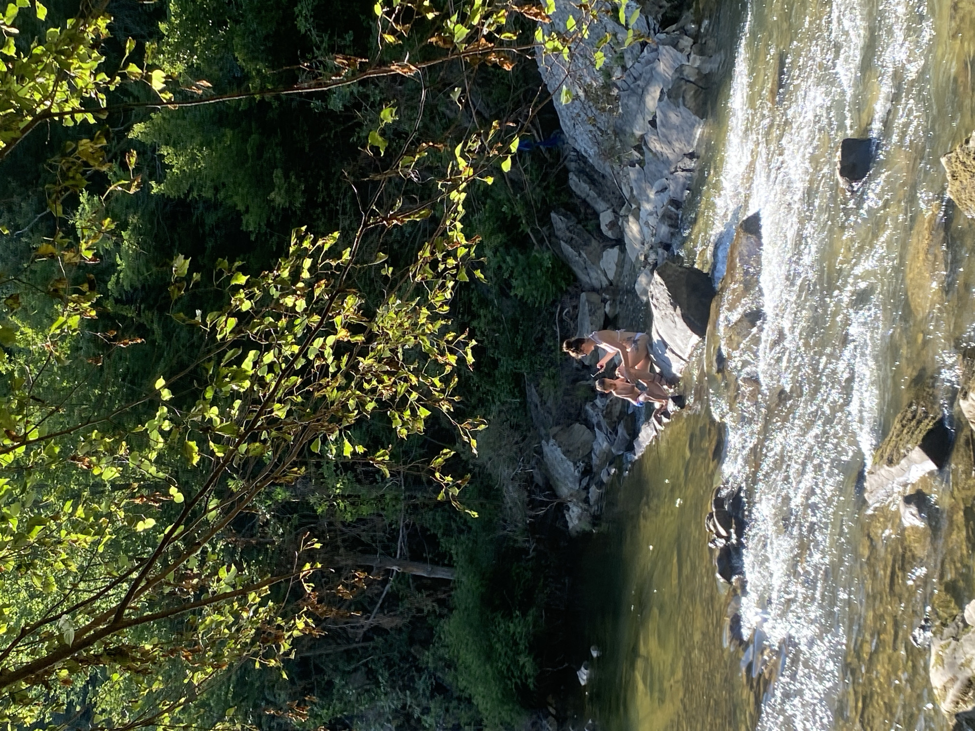 In Debnevo, Bulgaria, a woman and a young boy are sitting on a large rock in the middle of a shallow, clear river. The woman is wearing a light-colored swimsuit, and the boy is wearing blue swim trunks. They appear to be relaxed, with the woman looking down at her phone. The river is surrounded by lush green trees and large rocks. The water is flowing, creating small rapids around the rock where they are sitting. The sun is shining brightly, dappling the water and the surrounding foliage. The overall scene evokes a sense of peacefulness and enjoyment of nature.