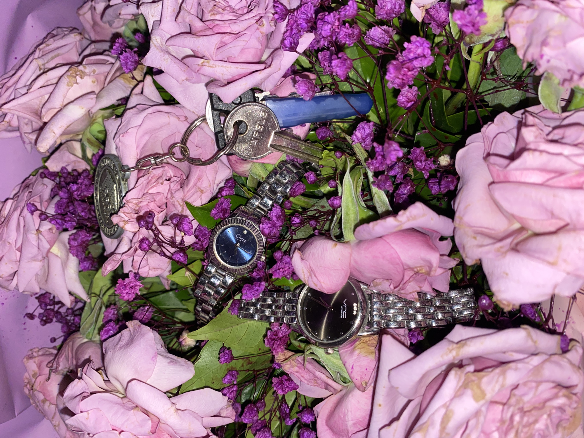 The image depicts a bouquet of light purple roses and small purple flowers. Scattered among the flowers are two silver-banded wristwatches, one with a black face and one with a blue face. A set of keys, including a blue-handled key and a silver key with a keychain, is also present among the floral arrangement. The location in the city of Davao City, Philippines, is not discernible from the image.