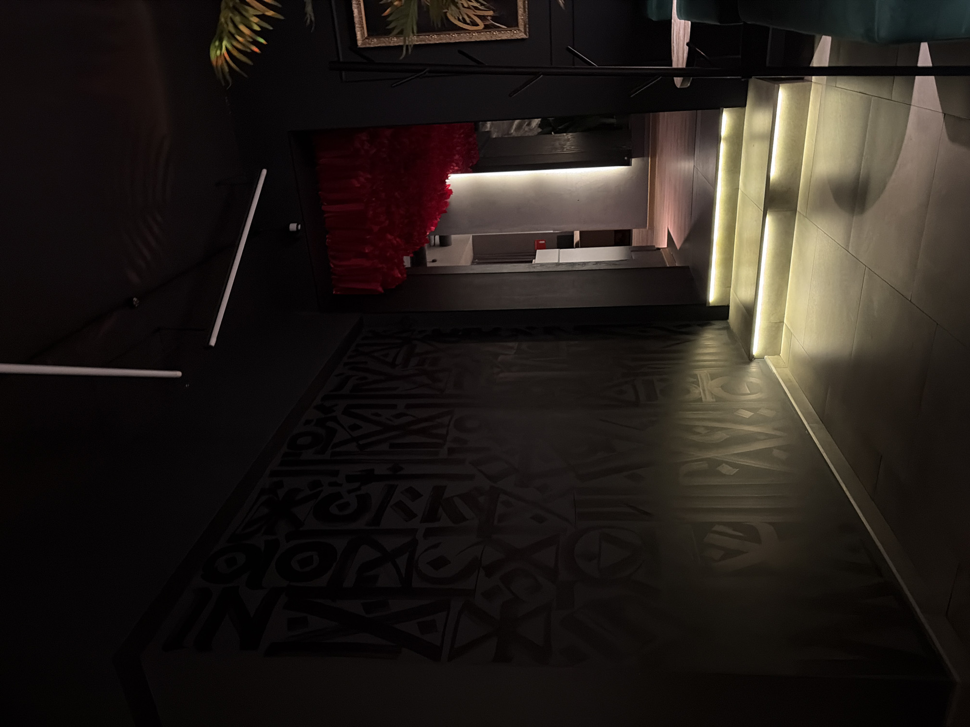 The image depicts the interior of a building in Chaikovsky, Russia. The location has a dark, modern aesthetic with distinctive lighting and design elements. A section of the floor is covered with abstract, dark-colored patterns. The architectural layout reveals an entryway or passage with illuminated stairs on the right side. A red, decorative element is suspended from the ceiling. A piece of art or foliage is partially visible in the upper portion of the frame.