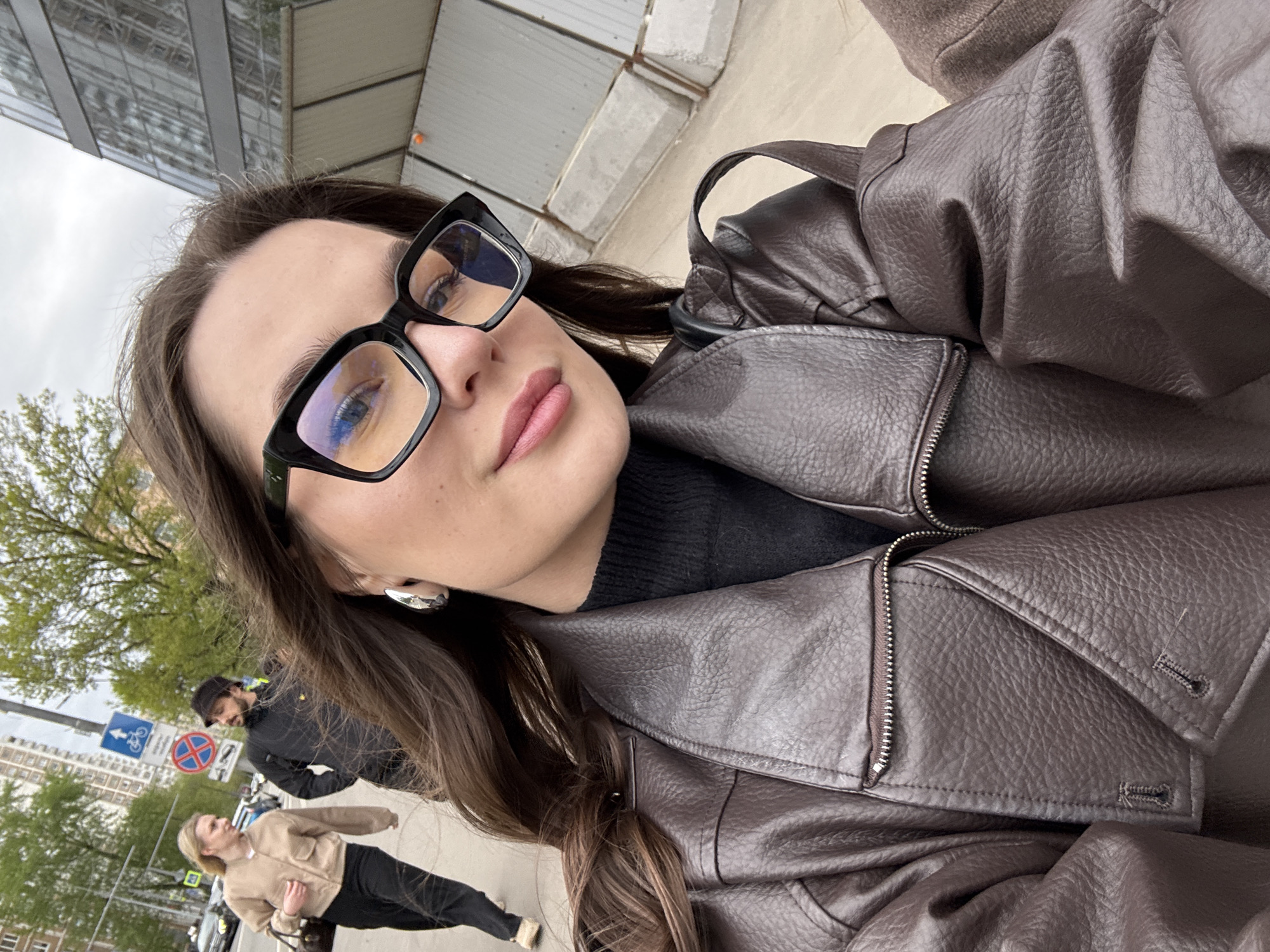 The image is a selfie of a woman wearing black-framed glasses and a brown leather jacket. She is standing on a sidewalk, with a city street and buildings in the background. Two other people can be seen in the background walking down the sidewalk. A bike path sign and a building with green trees in front of it are also visible. Based on architectural styles and the bike path sign, the location is likely in Moscow, Russia.
