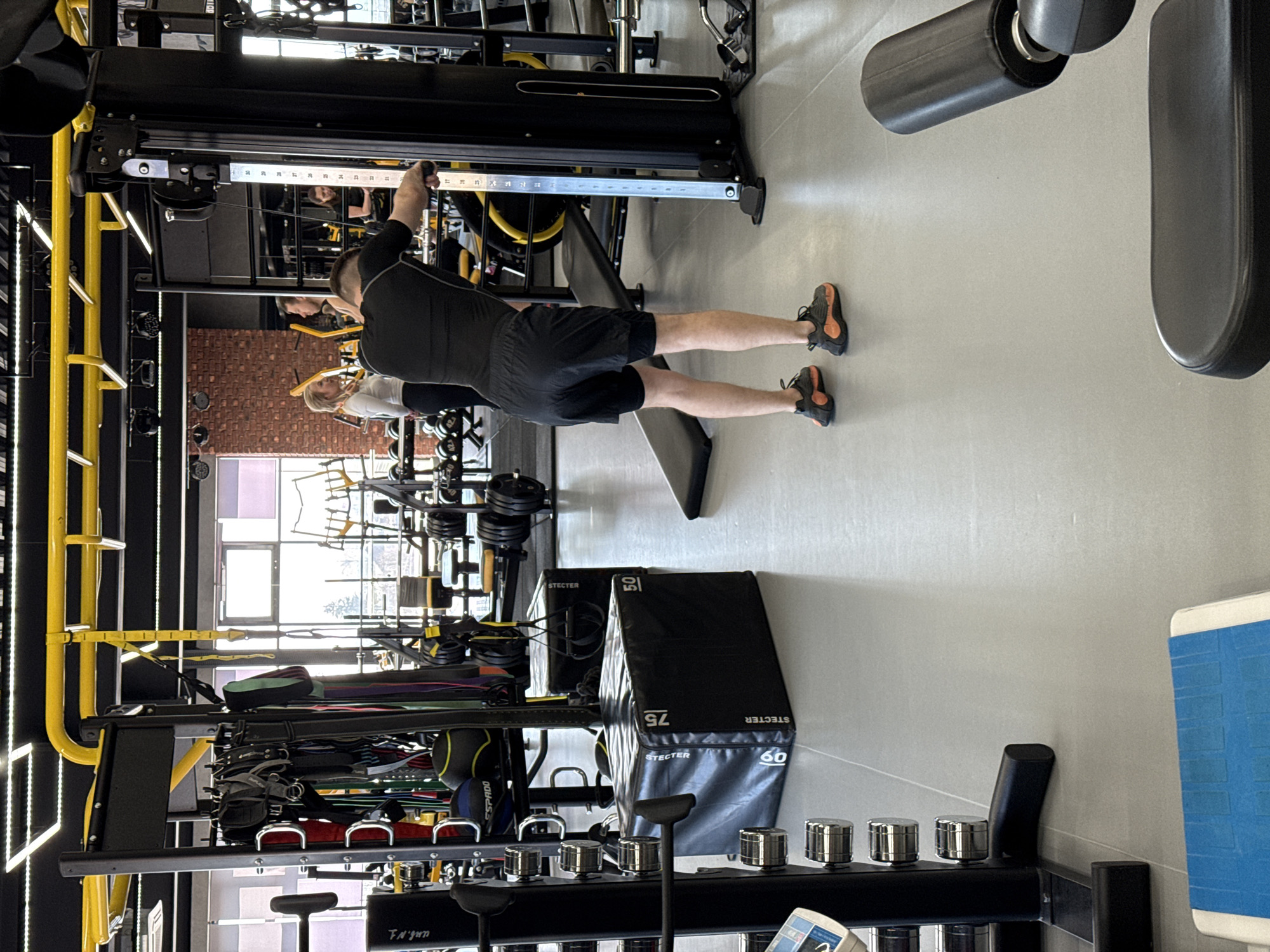 A man in black athletic wear performs a standing chest fly exercise at a modern gym in Votkinsk, Russia. The gym is well-equipped with a variety of fitness machines and equipment. The man faces towards the left, gripping the cable handles of a weight machine. Other gym members are visible in the background. The floor is light gray, and the gym appears to be well-lit, with natural light coming through the windows. Weight stacks, storage containers, and exercise accessories are arranged in the space.