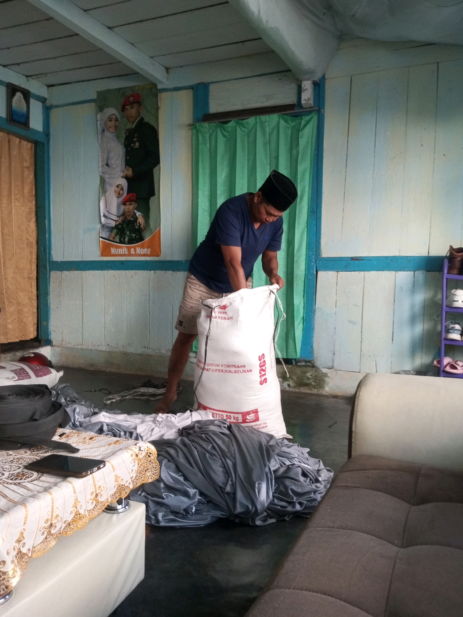 A man wearing a blue t-shirt and a black cap is opening a large white sack labeled "S12GS". The sack appears to contain some sort of powder or granular material, and text on the sack indicates it is 50 kg. The man is standing barefoot on a dark floor inside a room. The room has light blue and white wooden walls and a ceiling with exposed beams. A large green curtain hangs on the right side of the room, and a framed picture of a couple in military uniforms is on the back wall, with smaller figures below them. There are also stacks of folded gray fabric in the foreground. A table with a patterned tablecloth and a black smartphone is visible on the left. A brown sofa is on the right. The scene appears to be an indoor storage or living area in Andongsili, Indonesia.