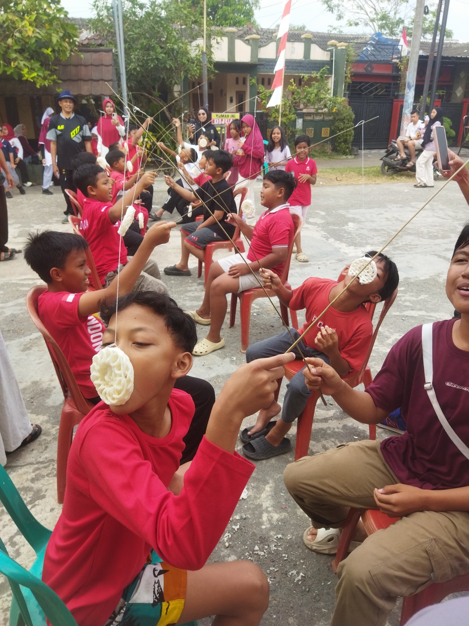 The image depicts an outdoor game likely in Banjar, Indonesia, as evidenced by the red and white Indonesian flag decorations visible in the background. Several children, mostly dressed in red shirts, are seated on plastic chairs. They are participating in a game where a circular cracker is suspended by a string in front of their faces, and they are attempting to eat it without using their hands. The children's facial expressions suggest concentration and effort. In the background, adults and other children are observing, and some are taking pictures. The environment appears to be a street or courtyard with houses and trees.