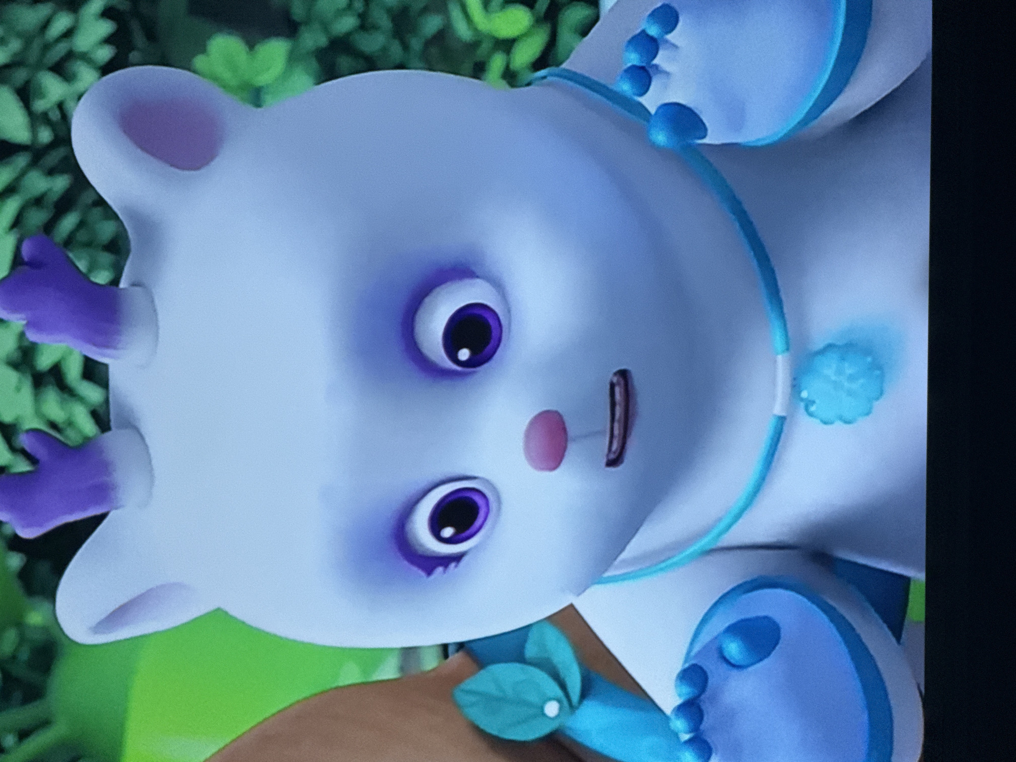 A close-up shot shows a light blue cartoon-style animal figure with purple accents around the eyes and on its small antlers. The figure has large, round purple eyes and a pink nose. It is wearing a light blue collar with a decorative pendant and a blue band around its body. The background is blurred, showing green foliage. The location is not discernible.