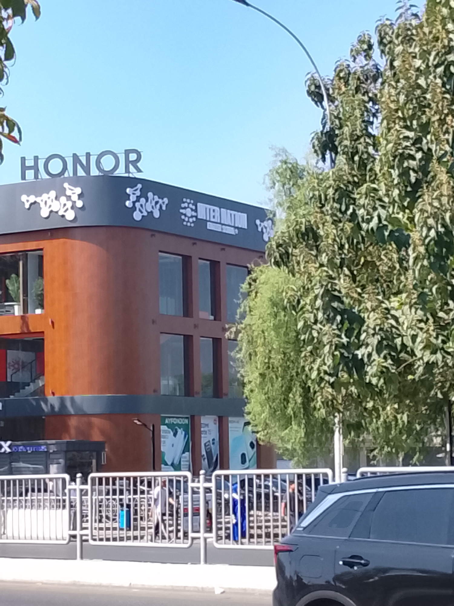 The image shows a building with the "HONOR" logo on its facade, located in Fergana City, Uzbekistan. The building has a modern architectural style, with a prominent curved brown exterior and large glass windows. The facade is decorated with white abstract designs. Trees with green foliage are visible in the foreground and to the right of the building. A white metal fence runs along the front of the property, and a dark-colored SUV is parked in front of it. Several people are visible behind the fence, some walking and others standing. The sky is clear and blue. The scene appears to be captured during daylight hours.