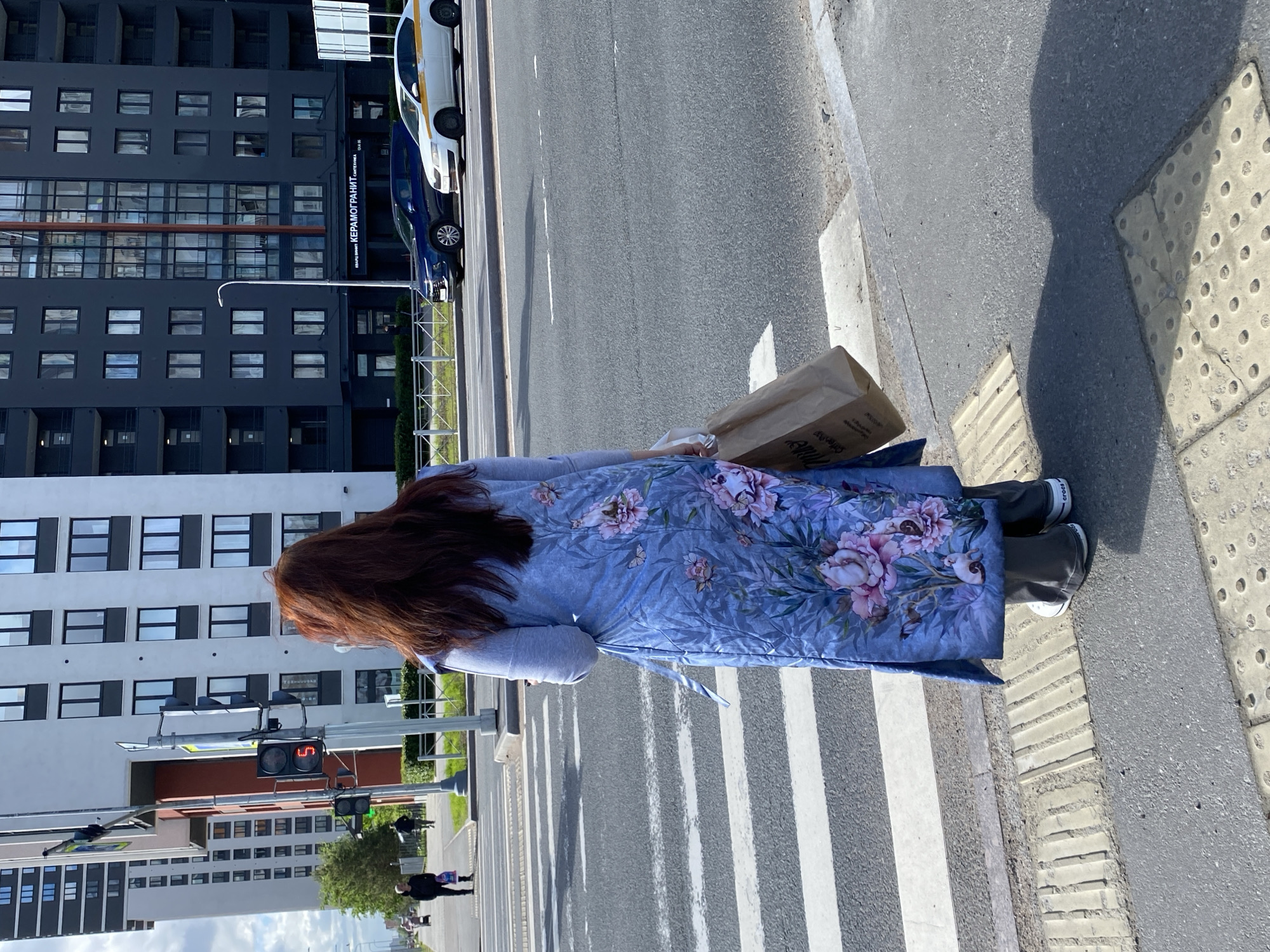 A woman with long, auburn hair is standing on a crosswalk in what appears to be Saint Petersburg, Russia. She is wearing a long, floral-patterned robe over a grey top. She is holding a brown paper bag with lettering in Cyrillic, possibly from a cafe or store. The crosswalk has white stripes on a grey asphalt road. A building with many windows is on the left side of the frame, and vehicles are parked on the right. In the background, traffic signals are visible. The tactile paving for pedestrian crossings is visible on the sidewalk.