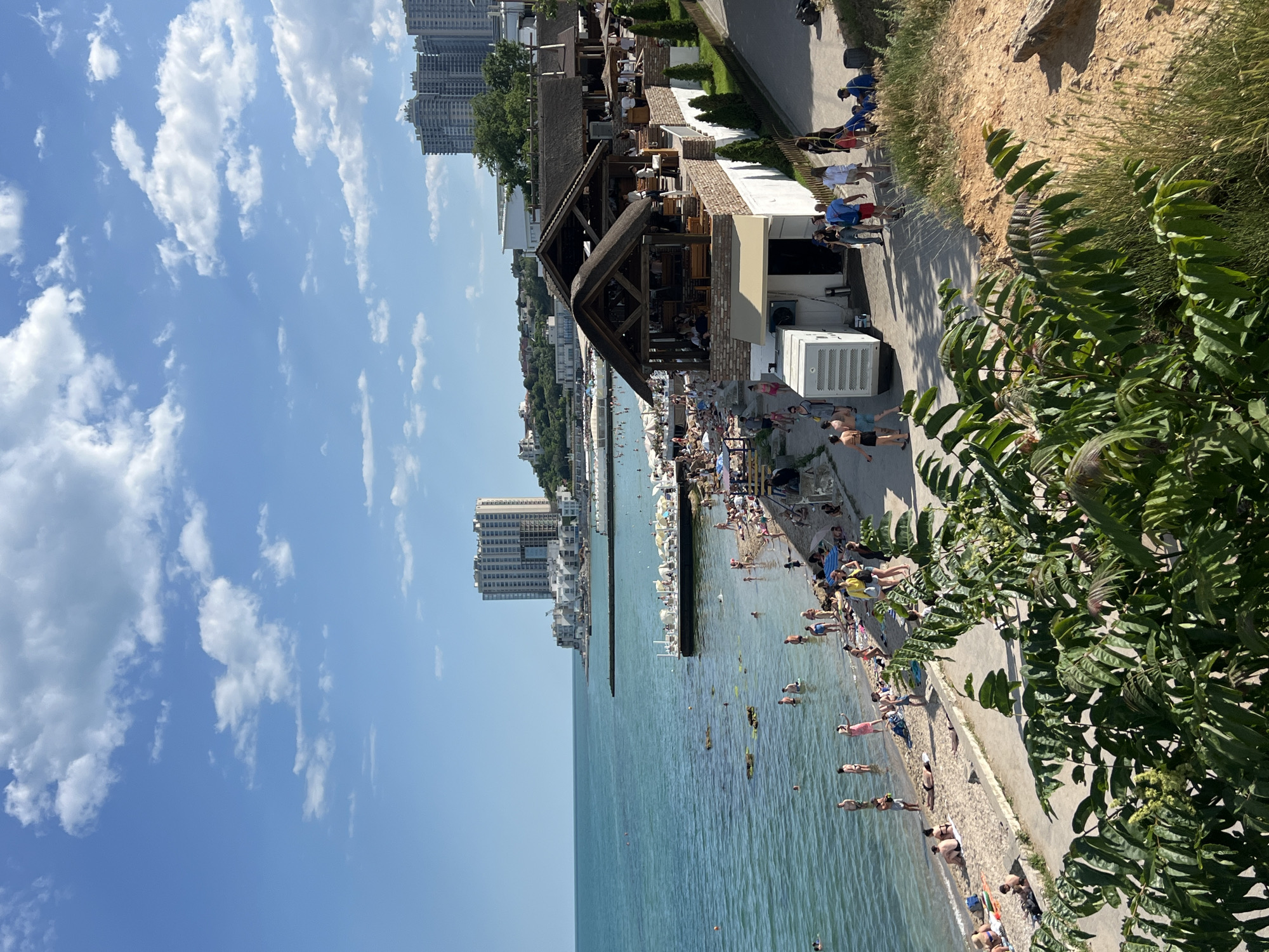 The image depicts a vibrant summer day at a coastal area in Odesa, Ukraine, likely near Arcadia Beach, characterized by its popularity for leisure and entertainment. A bright blue sky with scattered white clouds stretches across the upper portion of the frame. Below, the tranquil, turquoise waters of the Black Sea meet a sandy beach teeming with activity.
Numerous people are enjoying the sun and surf. Some are wading in the shallow water, while others are sunbathing on the sand, relaxing on beach chairs, or strolling along the shoreline. Beach umbrellas provide shade for some of the sunbathers.
Adjacent to the beach is a bustling promenade lined with a restaurant or cafe featuring thatched roofs and open-air seating. Several individuals are visible dining or socializing at the establishment. A white, portable generator unit is positioned near the entrance of the building. A paved walkway runs alongside the beach and the restaurant, with people walking in both directions.
Further inland, and in the background, the distinctive architecture of Odesa is visible, with several tall, multi-story residential buildings rising against the skyline. Greenery, including trees and shrubs, frames the scene, adding to the picturesque coastal ambiance. The overall impression is one of a lively and enjoyable summer day at a popular seaside resort.