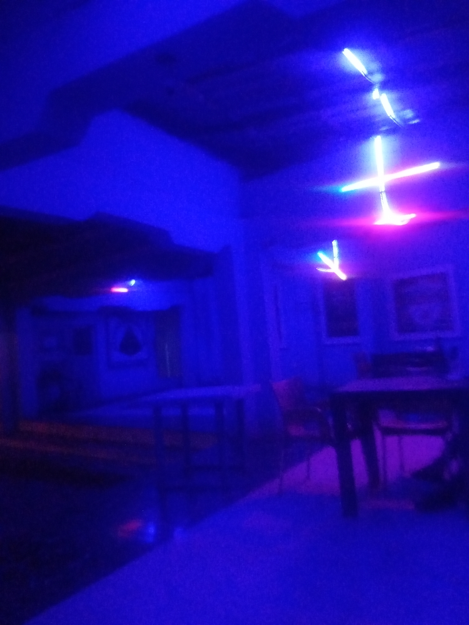 The image shows a dimly lit interior space, likely a room or hall, dominated by deep blue and purple lighting. Several illuminated neon or LED light fixtures are visible, some shaped like crosses or arrows, suspended from the ceiling. In the foreground, a dark, indistinct table and chairs are present. The background features what appear to be framed pictures or decorative elements on the walls, also bathed in the pervasive blue light. The overall impression is one of a space with a deliberate, atmospheric lighting design, potentially a club, bar, or art installation. The location of Sabon Gari Nassarawa, Nigeria, cannot be determined from the visual information in the image.
