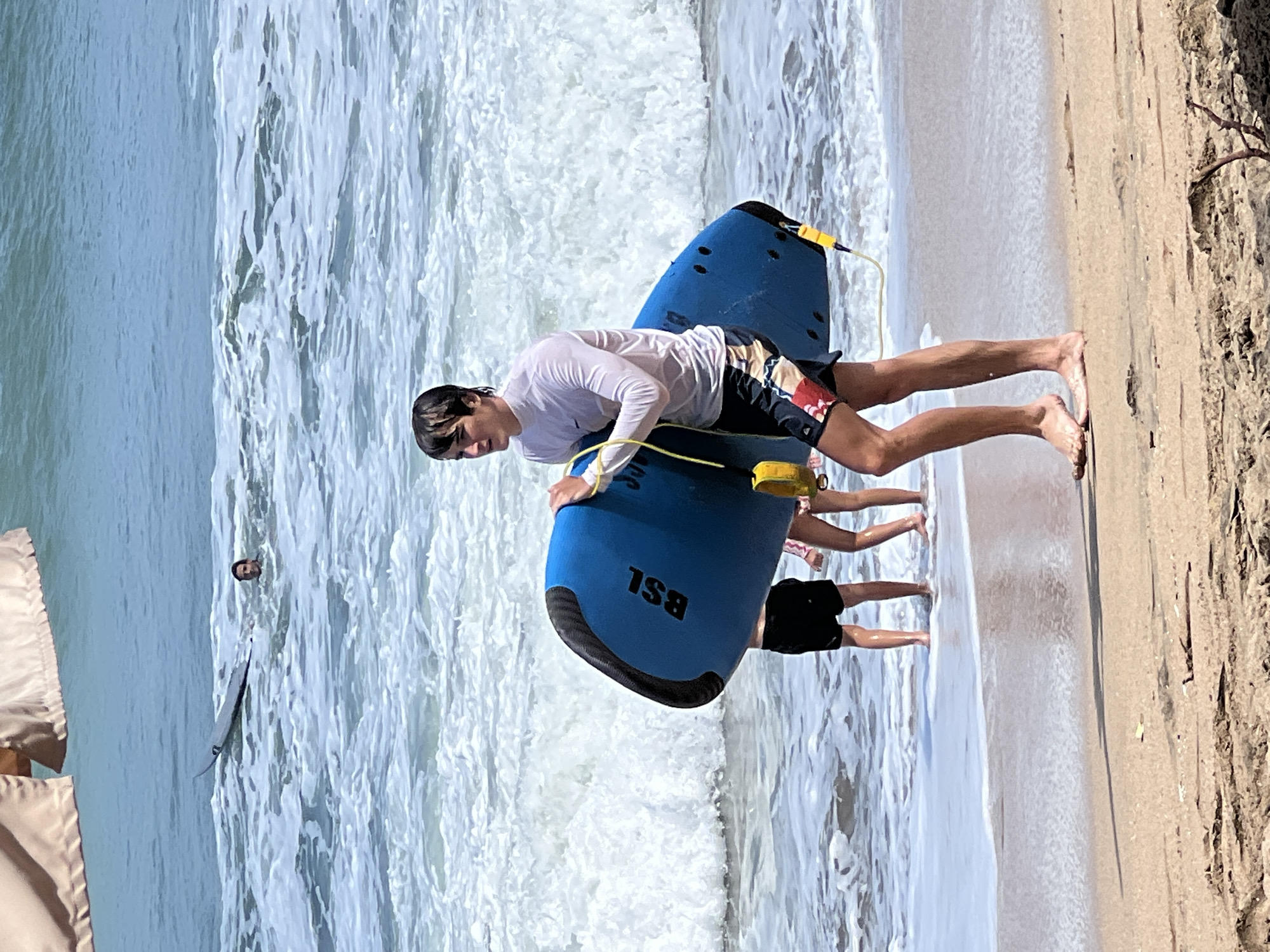 A young man, likely a tourist in Kuta, Indonesia, walks on a sandy beach carrying a blue boogie board. He is wearing a white long-sleeved rash guard and patterned swim shorts, his hair wet from the ocean. The waves are breaking on the shore behind him. Two children, partially obscured by the boogie board, stand in the shallow water, their legs visible. Further out in the ocean, another person is visible in the water, near a surfboard. The sky is clear and blue, and the sun is bright. In the foreground, some dry seaweed and driftwood are scattered on the sand. The overall scene depicts a typical beach day at a popular tourist destination like Kuta.