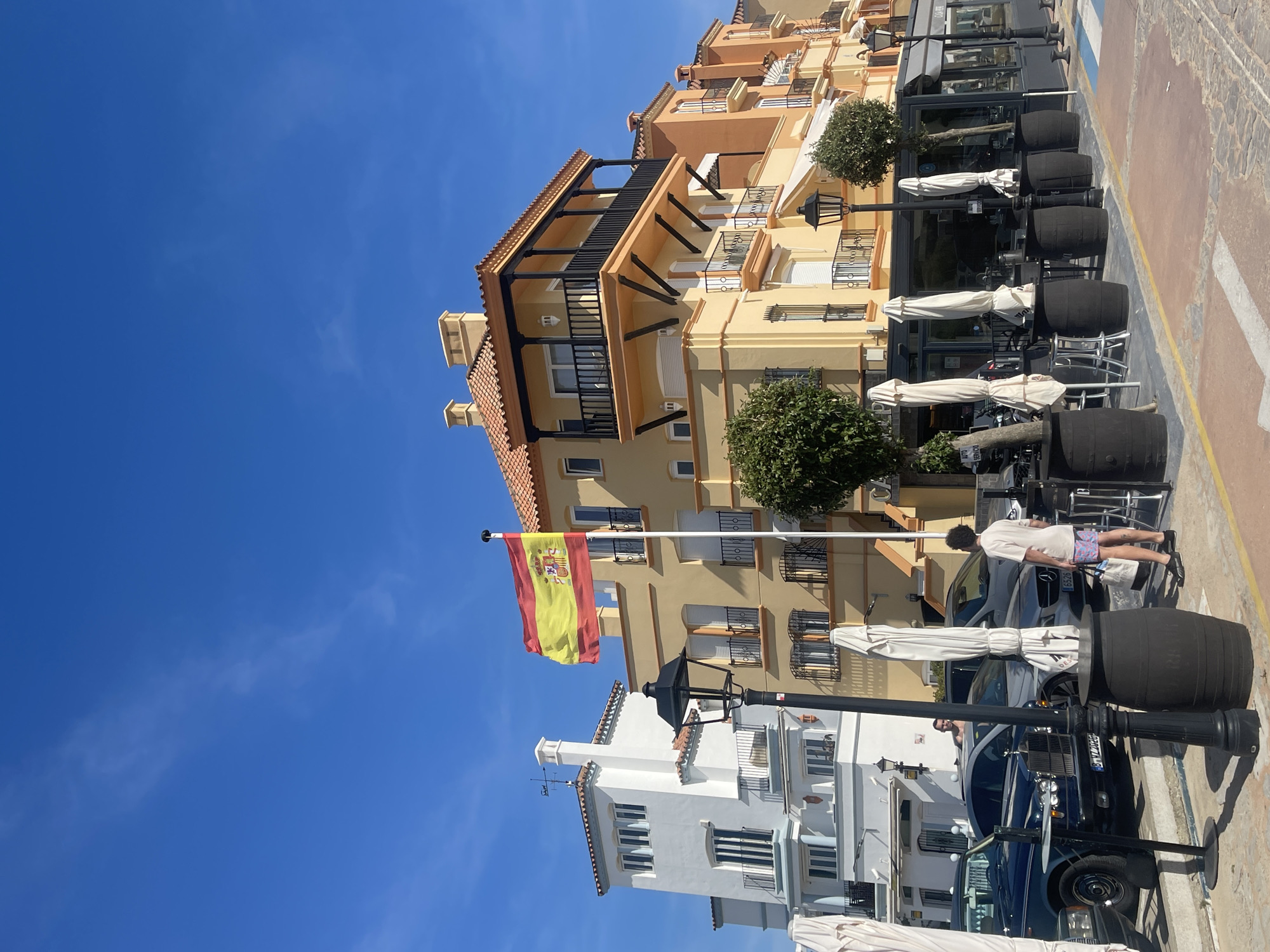 The image shows a street scene in El Puerto de Santa María, Spain. The foreground features a row of barrels likely repurposed as tables or decor for a restaurant or café. Umbrellas are folded and positioned near these barrels. Several cars are parked along the street. A person is walking between the vehicles and the barrel arrangement. The Spanish flag is prominently displayed from a pole attached to a building. The buildings in the background are of varying heights and architectural styles, with one white building contrasted against a multi-story yellow building with balconies. The sky is clear and blue.