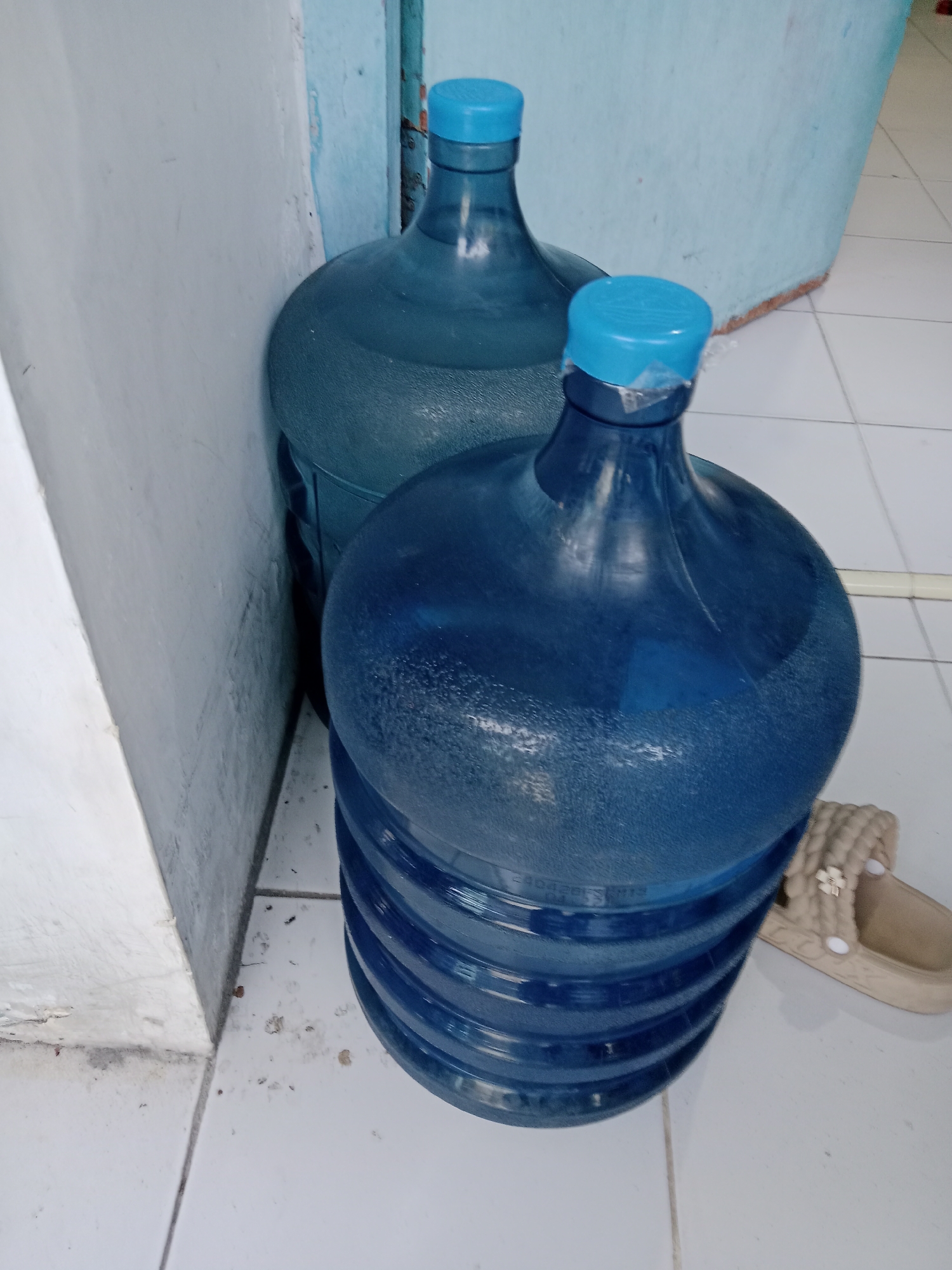 The image shows two large blue plastic water jugs with blue caps, standing in a corner next to a light-colored wall. The jug in the foreground is larger and appears to be full, with visible condensation on its surface. The jug behind it is slightly smaller and partially obscured. Both jugs have ridged bases. To the right of the foreground jug, a beige sandal with a braided strap is visible. The floor is tiled in white. The environment appears to be indoors, possibly a home or storage area. The scene is static and shows no activity.
