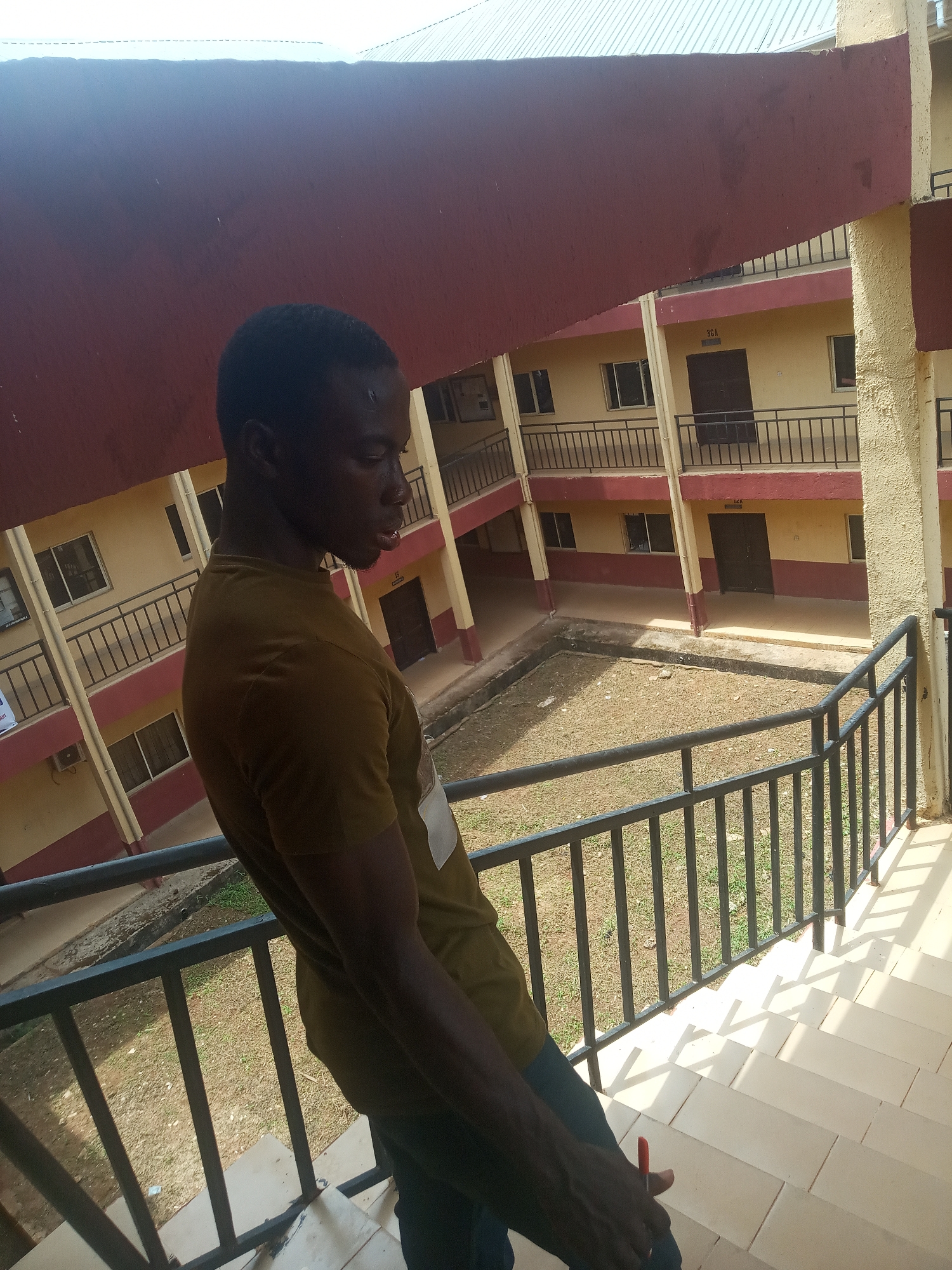A young man, seen in profile, stands on an upper floor balcony, gazing downwards into a central courtyard. He has dark skin, short dark hair, and is wearing a dark olive/brown short-sleeved t-shirt and dark trousers. A small red object, possibly a pen, is visible in his right hand near his hip.
The setting is a multi-story building, likely an institutional or residential complex in Nkwelle, Nigeria. The architecture features light yellow walls accented with maroon/red on the balcony edges and support pillars. Each floor has a black metal railing. In the background, numerous windows and dark doors are visible along the corridors overlooking the courtyard.
The courtyard below is a patch of dry earth with sparse green growth and concrete borders. Strong, bright sunlight illuminates the scene, casting sharp shadows from the railings onto the light-colored tiled floor where the man is standing, suggesting it is daytime.
Visible text includes room or block identifiers such as "1C1 BLOCK A" (partially visible), "1CA", and "15" on the building walls in the background. The man's posture appears contemplative or observant.