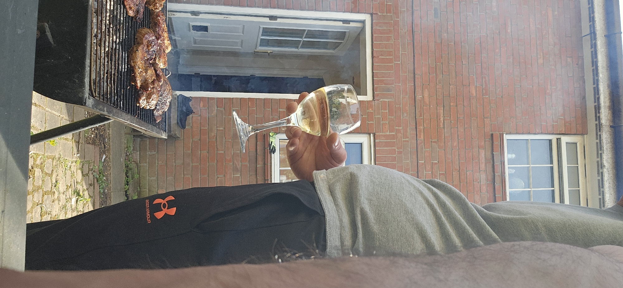 A point-of-view shot shows a person reclining, holding a wine glass filled with a light-colored liquid. A barbecue grill laden with grilled meat is visible to the left. In the background, there's a brick building with two windows, one open. The person wears black Under Armour pants and a gray shirt. A shoe is sitting on the grill behind the grill meat. The location is indeterminate and cannot be verified with this image.