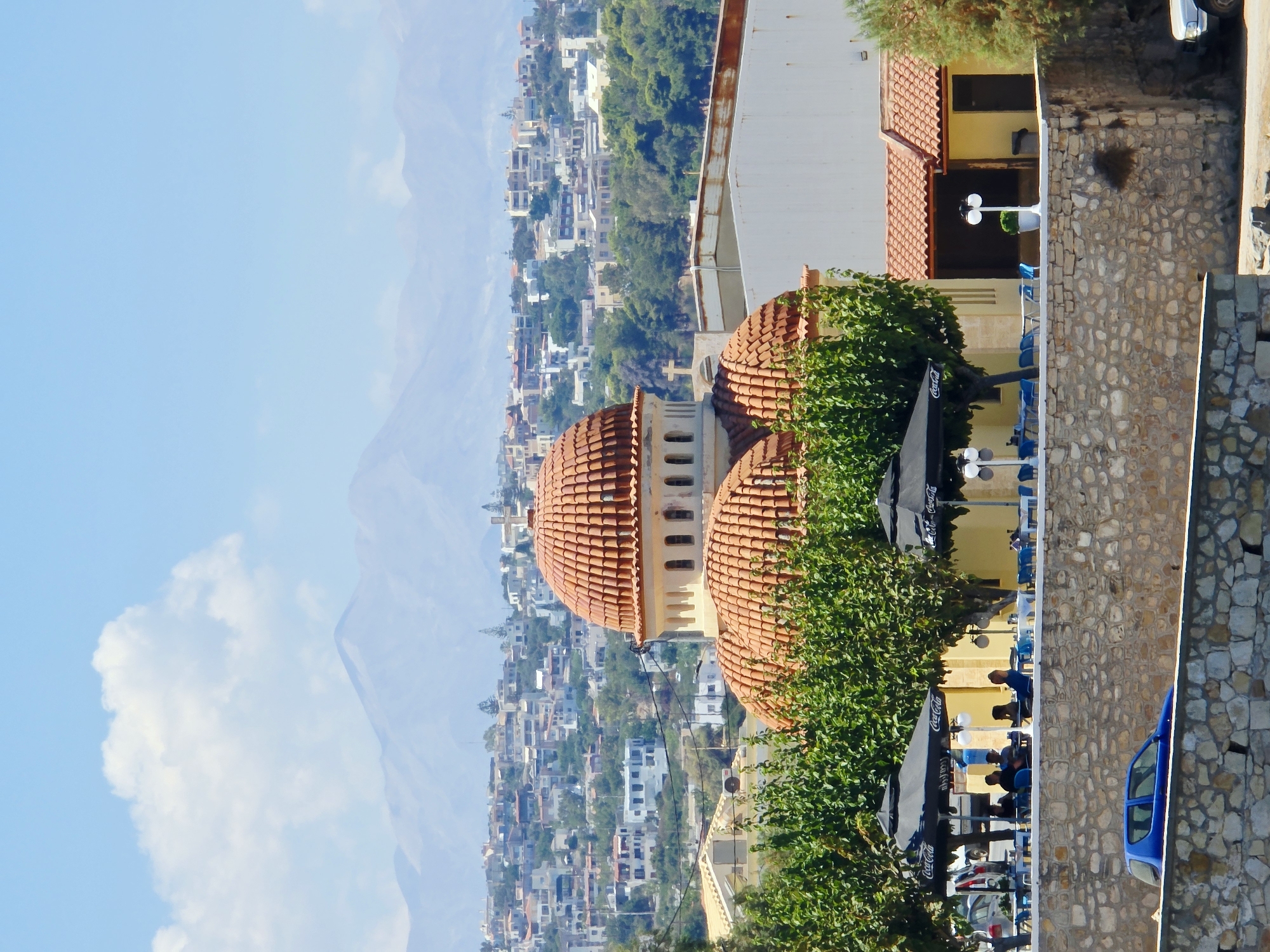The image shows a picturesque view of Rethymno, Greece, with a prominent domed building in the foreground. The building features a traditional terracotta tiled roof and appears to be a church or similar structure, with a cross visible on its roof. Behind the building, a densely populated city stretches across rolling hills, characterized by white and pastel-colored buildings. In the distance, imposing mountains form a backdrop against a clear blue sky with scattered white clouds.
In the immediate foreground, a stone wall separates the viewer from a lively outdoor seating area of a cafe or restaurant. Several tables are set up under black umbrellas, some with the Coca-Cola logo visible. People are seated at some of the tables, engaged in conversation or enjoying their meals. A blue car is parked on the street behind the stone wall, and further to the right, another light-colored vehicle is partially visible. Lush green trees provide shade and add to the vibrant atmosphere. The overall scene suggests a warm, sunny day in a charming coastal town.