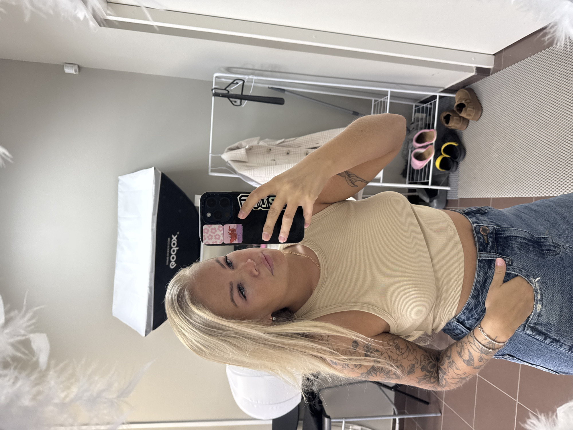The image displays a mirror selfie of a young woman taken indoors, presented on its side. When viewed upright, the woman is seen from the waist up, facing the mirror and holding a black smartphone with a case decorated with two small stickers – one with pink flowers, the other with a red crab or lobster – and what appears to be stylized text like "FDUG". She has long blonde hair, which falls over her left shoulder and arm. She is wearing a light beige ribbed tank top and blue denim jeans, with her right hand tucked into her jeans pocket. Numerous tattoos, including floral designs and what looks like a bird or fish, cover both of her arms. Her expression is neutral, and she is looking directly into the phone's camera.
The background reflects an indoor setting. To the left of the woman, a white softbox light source is visible, which has text printed on it that reads "qobox" and "www.qobox.com" (likely reversed from "Godox"). Behind her, there is a white metal clothing rack with a light-colored, possibly plaid, shirt hanging on it. Below the clothing rack, several pairs of shoes are placed on the floor, including brown slippers and pink and yellow slip-on sandals. The floor transitions from a light-colored, textured mat to a brown tiled section. The walls are a plain light grey or beige. A small white cylindrical object is mounted on the wall or ceiling in the upper background.