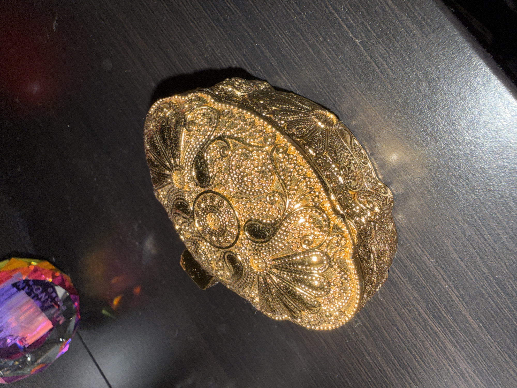 A highly ornate, gold-colored, oval-shaped trinket box sits on a dark, textured surface that appears to be wood grain. The box is intricately detailed with raised floral and swirling patterns, and small textured dots that catch the light. The surface beneath the box has diagonal lines, suggesting a brushed or textured wood grain. In the upper left corner of the image, a faceted, multi-colored glass object, possibly a paperweight, reflects light in shades of purple, pink, and yellow. This object also has writing on it, with the word "MILANO" being partially visible. The lighting in the scene is dramatic, with bright highlights on the gold box and the glass object, and deep shadows. The overall impression is of a still life arrangement on a dark surface, possibly a tabletop. The location in Pamplona, Spain is not ascertainable from the visual information present in the image.