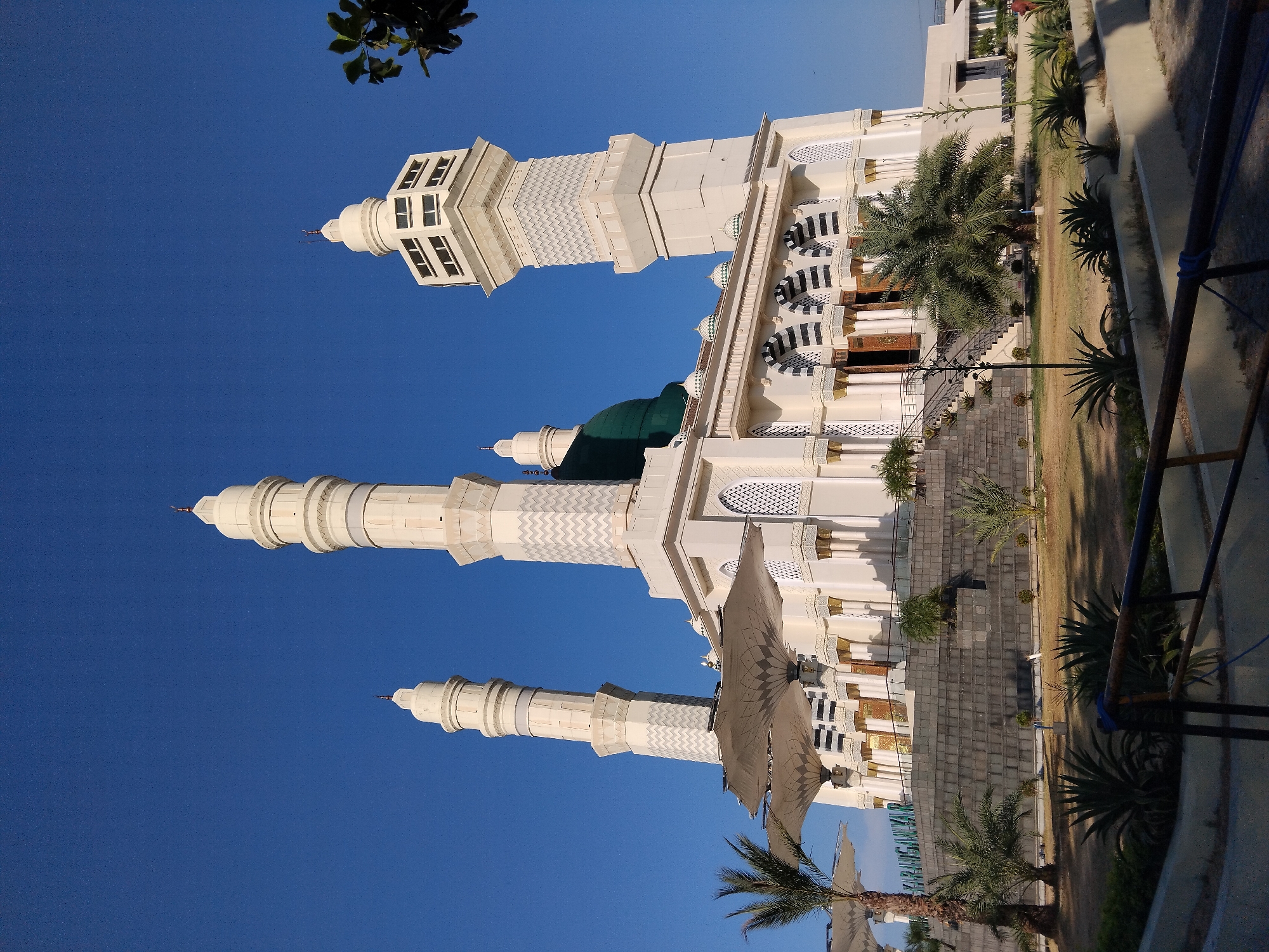 This is a low-angle, exterior, full shot of a mosque in Karanganyar, Indonesia, under a clear blue sky. The mosque features a central, large green dome and is flanked by three tall, white minarets. The minarets have a cylindrical base that tapers upwards, with decorative patterns and windows near their summits. The mosque's main structure is white with arched entrances and windows featuring intricate geometric designs. In front of the mosque, there are large, white, umbrella-like canopies supported by poles. The foreground shows a landscaped area with palm trees, other types of plants, and a pathway leading up to the mosque. A stone-tiered foundation leads to the mosque entrance, and a stone-paved area with railings is visible in the foreground. The word "KARANGANYAR" is visible in green lettering on the mosque's facade. No people are visible in the image. The lighting suggests it is daytime, with the sun casting shadows.