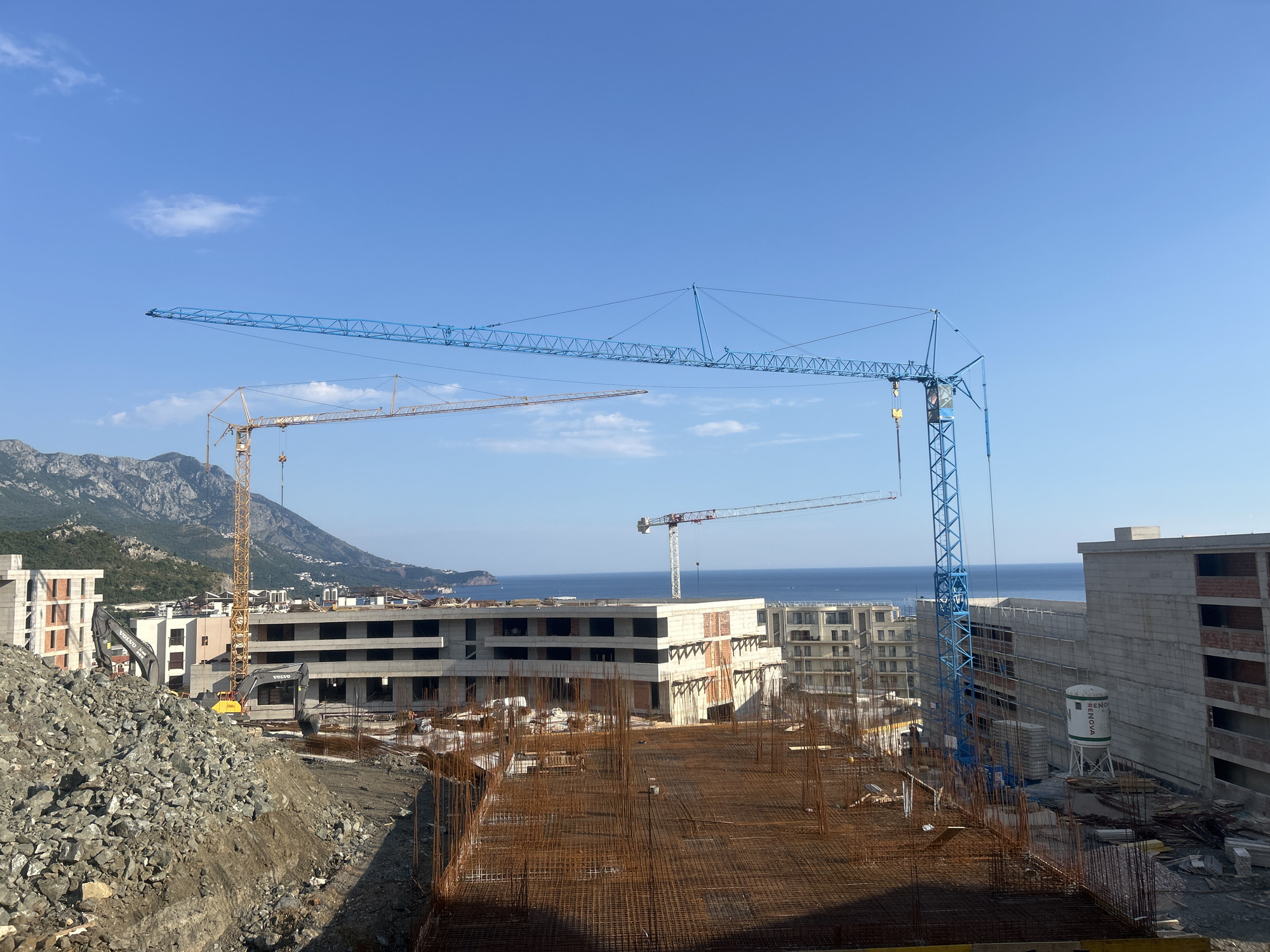 This media file depicts an outdoor construction site on a clear, sunny day, likely in the morning or early afternoon, given the bright light. The scene is dominated by several large tower cranes, one prominently blue and others in yellow and white, actively engaged in construction work. In the foreground, a substantial pile of grey rocks and dirt is visible, suggesting excavation or material staging.
Beyond the immediate construction area, unfinished buildings with exposed concrete and rebar structures are evident. Some buildings appear to have multiple stories, with visible window openings. In the background, a body of water, likely the sea, stretches to the horizon, bordered by lush, mountainous terrain. The sea is calm and reflects the blue of the sky.
Several construction vehicles, including two grey Volvo excavators, are present on the site. A white water tank with green text "RENOVA" is also visible. The overall impression is one of ongoing development and construction in a scenic coastal and mountainous region. The presence of multiple cranes and unfinished structures indicates a large-scale building project in progress. The clear sky and bright sunlight suggest favorable weather conditions for outdoor construction.