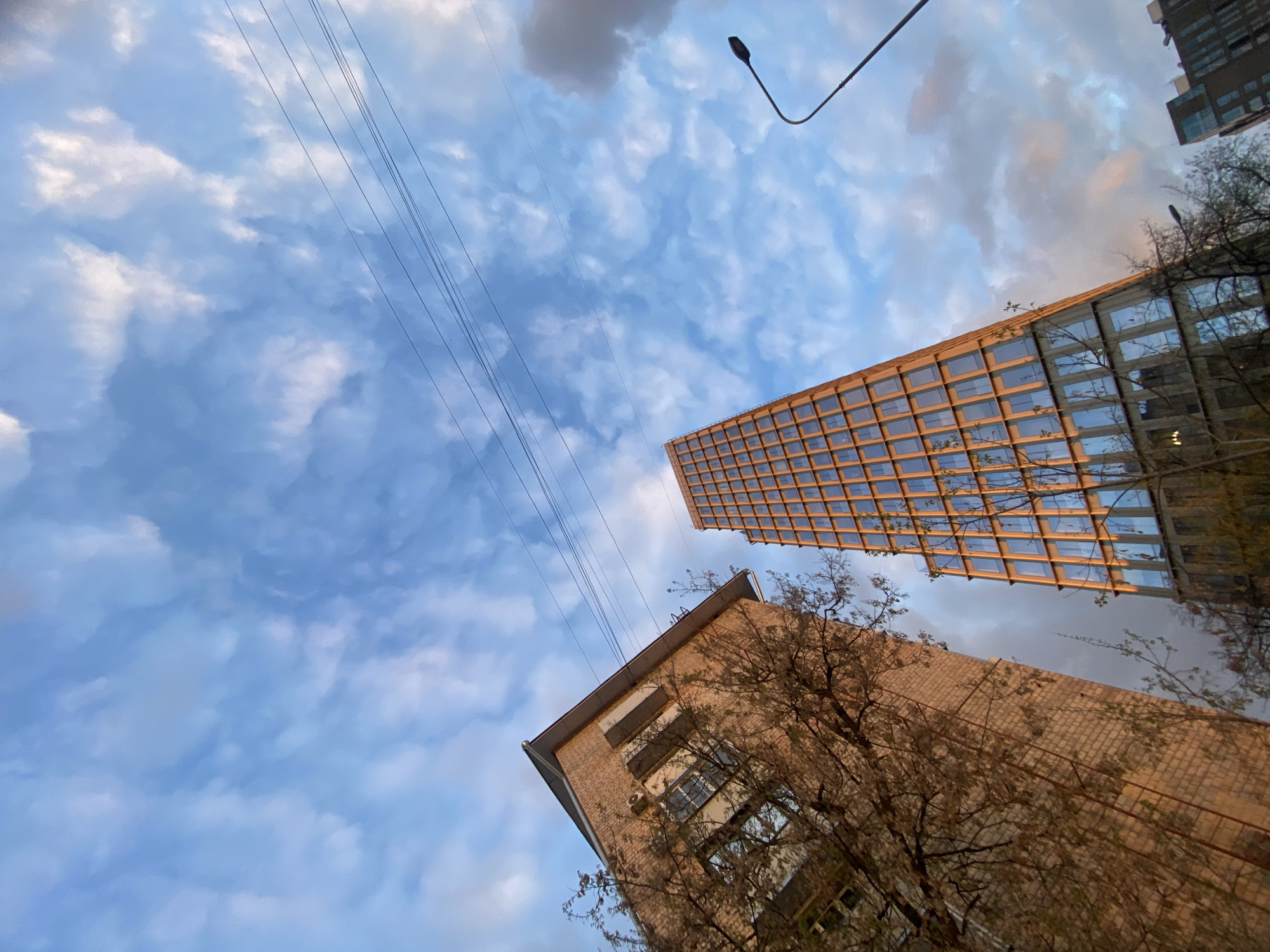 The image, taken from a low angle, shows two buildings and a cloudy sky with power lines. The building on the right is tall with many windows and has some foliage on its side. The other building is brick with fewer windows. A streetlight pole is in the upper right corner of the image. The sky is blue with white and gray clouds. Based on the architecture, the location is possibly in Moscow, Russia.