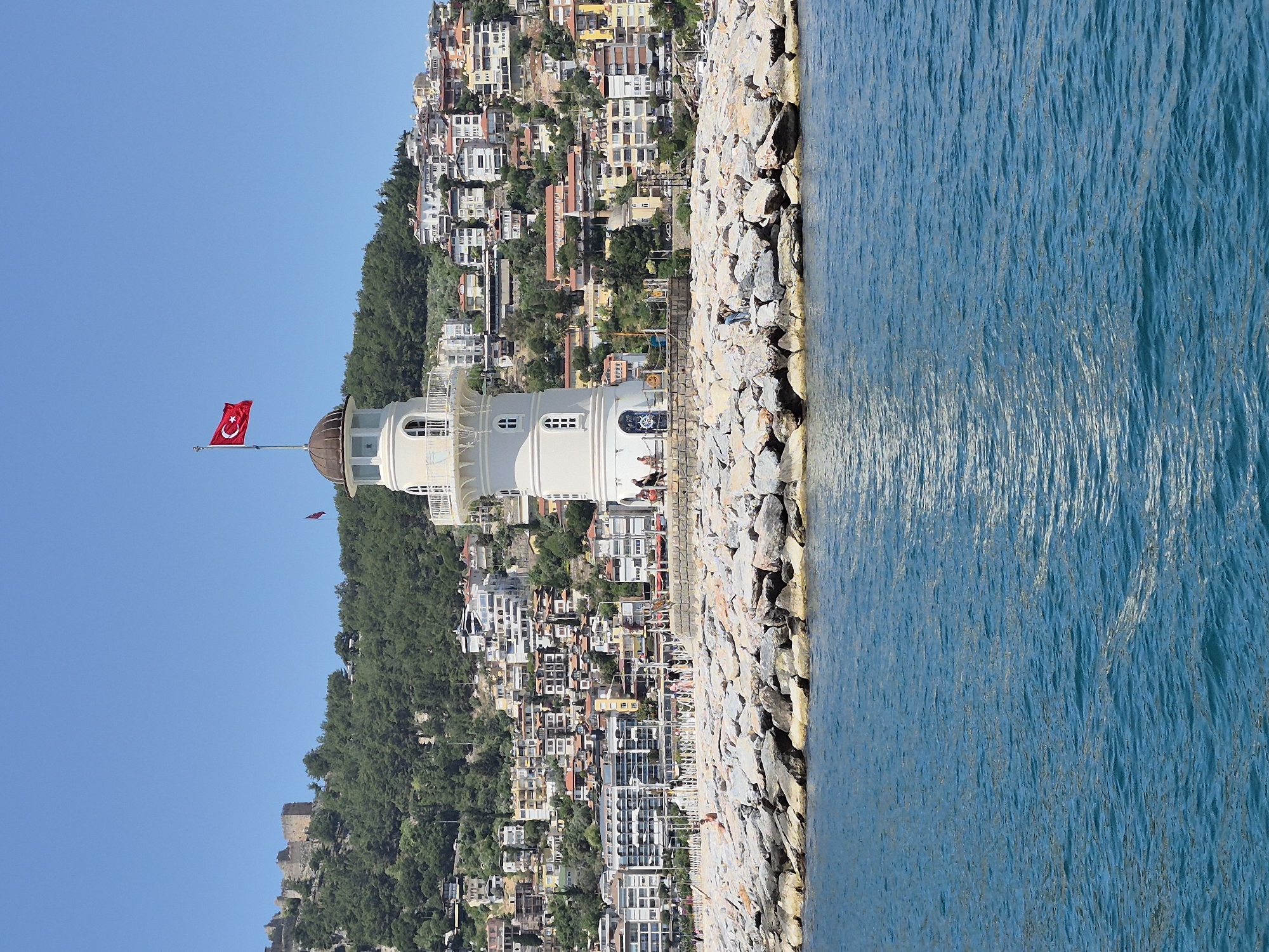 The image shows a lighthouse in Alanya, Turkey, standing on a stone breakwater. The lighthouse is white with a brown dome, and the Turkish flag flies from its top. People are visible on the breakwater near the lighthouse. The sea is to the right, and a hillside covered in buildings is behind the lighthouse. The sky is clear and blue. The overall scene is a sunny day at the coast.