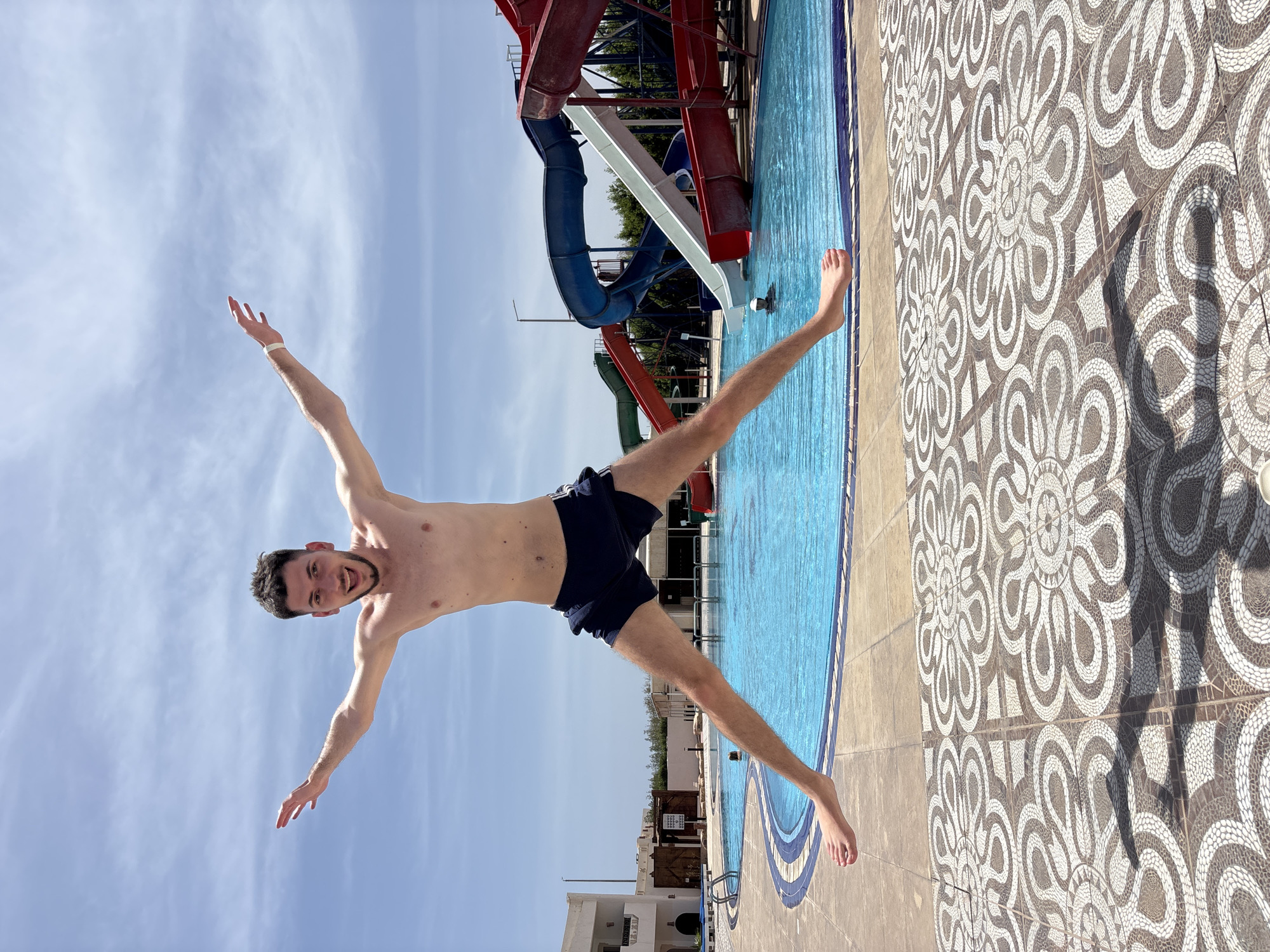 The image shows a fair-skinned man with dark hair and a beard, wearing dark swim trunks, in a cruciform pose with outstretched arms and legs. He is positioned at an angle, appearing to jump or fall. The backdrop includes a blue sky, a swimming pool with blue water, a water slide, and patterned tile flooring surrounding the pool. The city is Unknown, Unknown.