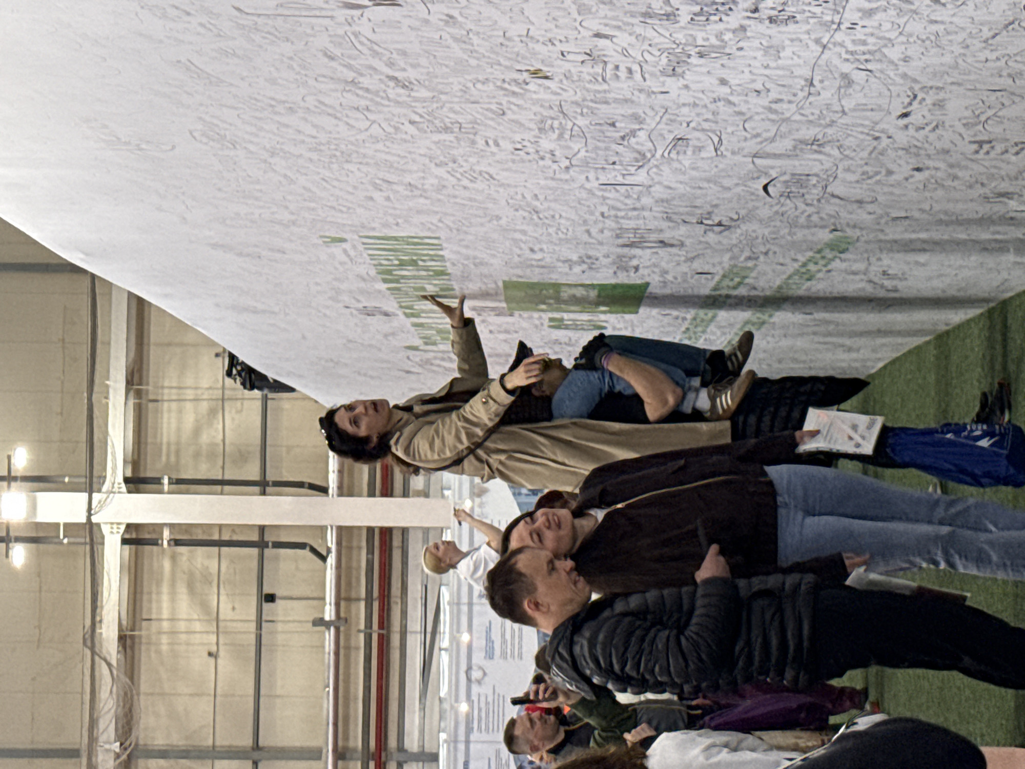 A low-angle, tilted-sideways shot depicts a group of people in a large indoor space, possibly a warehouse or convention hall, looking up at a large, off-white horizontal display. The display appears to be a large-scale map or drawing, featuring a detailed network of lines and some areas of green markings. A woman in a tan trench coat stands near the display with her arm raised. She is lying on her back. Below her, a second woman lies on her back. The third visible person is a man in a quilted black jacket. They are on an artificial turf ground. The building has light grey walls and metal beams. Other people are visible in the lower portion of the frame. Given the presence of the green markings and the description of the event, it is likely the location is an exhibition in Kazan, Russia, at the Kazan Expo, where the world's largest hand-drawn map was displayed in May 2024.