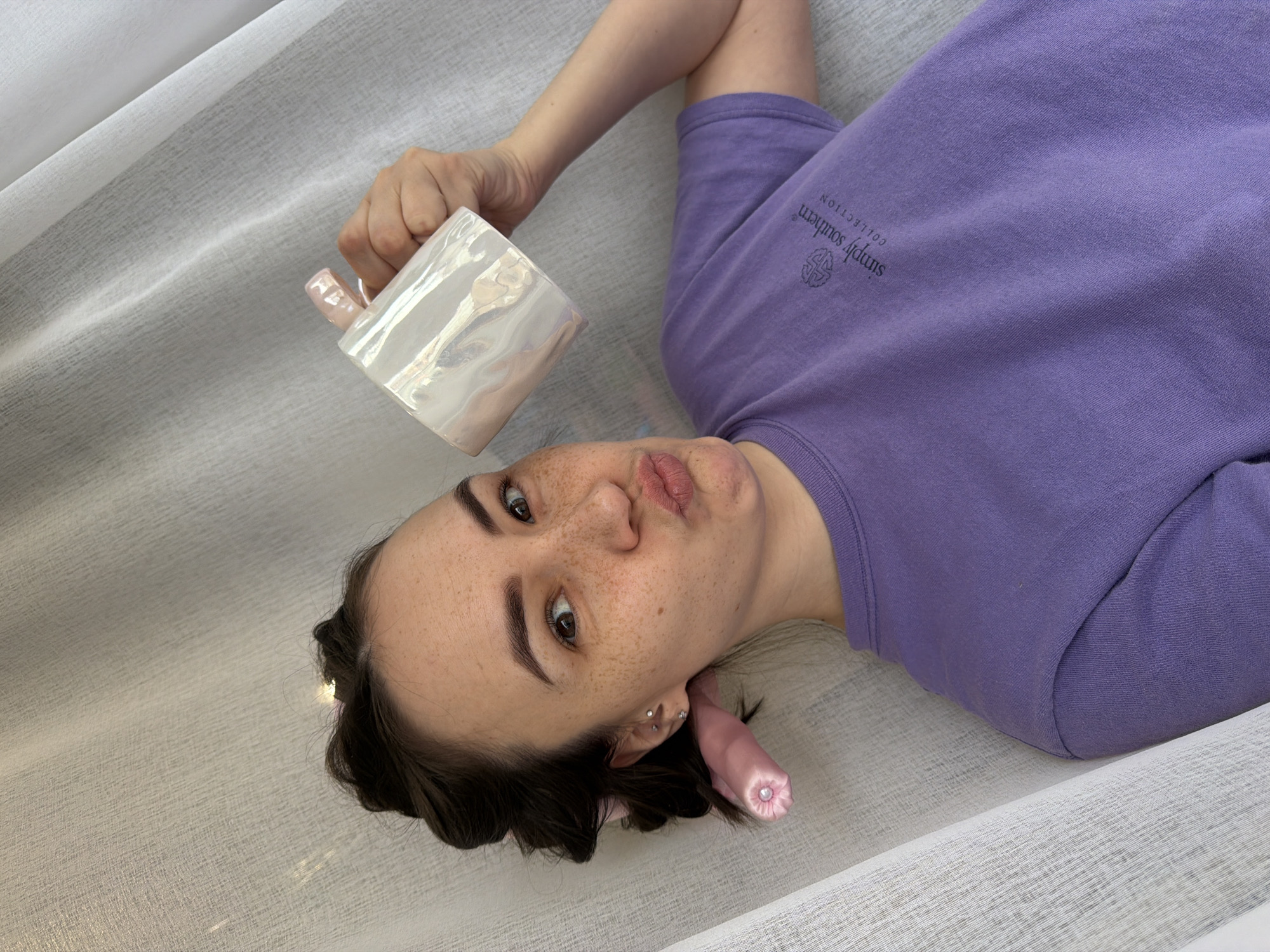 A woman is lying on a white, textured surface, possibly fabric. She is wearing a purple, long-sleeved shirt with the words "simply southern collection" printed on the upper right side. She is holding a white, iridescent mug. She has brown hair, light skin, and freckles. A pink object with a pearl-like embellishment is visible near her ear. The location is unspecified.