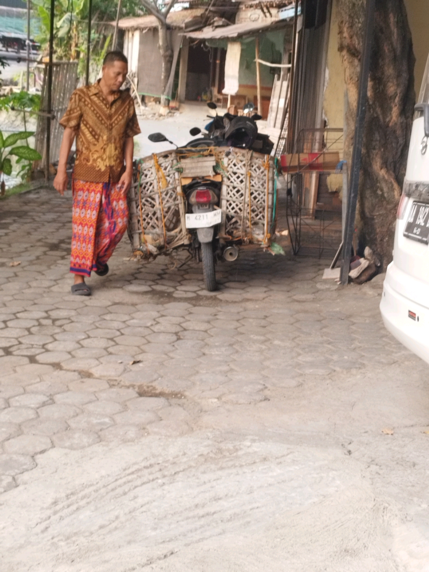 A man in traditional Indonesian clothing walks along a paved courtyard. He wears a patterned brown shirt and a vibrant orange sarong with a blue trim. His feet are clad in dark sandals. To his right, a motorcycle is parked, laden with a woven basket carrying what appears to be produce or goods. The motorcycle has a license plate reading "H 4211 MM". In the background, a building with a corrugated metal roof and wooden elements suggests a rural or semi-urban setting. A large tree trunk stands to the right of the motorcycle, and a white vehicle is partially visible further to the right. The ground is paved with hexagonal interlocking bricks. The scene appears to be in Patebon, Indonesia, based on the provided context.