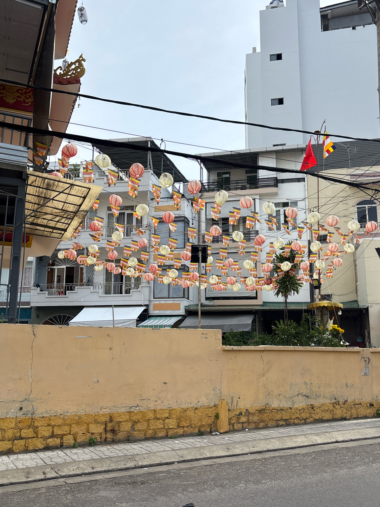 The image depicts a street scene in Nha Trang, Vietnam, likely near a religious site given the decorations. A line of red and white lanterns and Buddhist prayer flags are strung across the street, suspended by wires. These decorations are strung in front of several buildings, some of which are modern and white, while others appear to be older residential or commercial structures with balconies and awnings. A low, mustard-yellow wall with a yellow brick base runs along the bottom of the frame, separating the street from a sidewalk and the buildings beyond. The street itself is paved, with a concrete curb. The sky is overcast. There are no people visible in the frame. The overall impression is of a street decorated for a festival or observance.