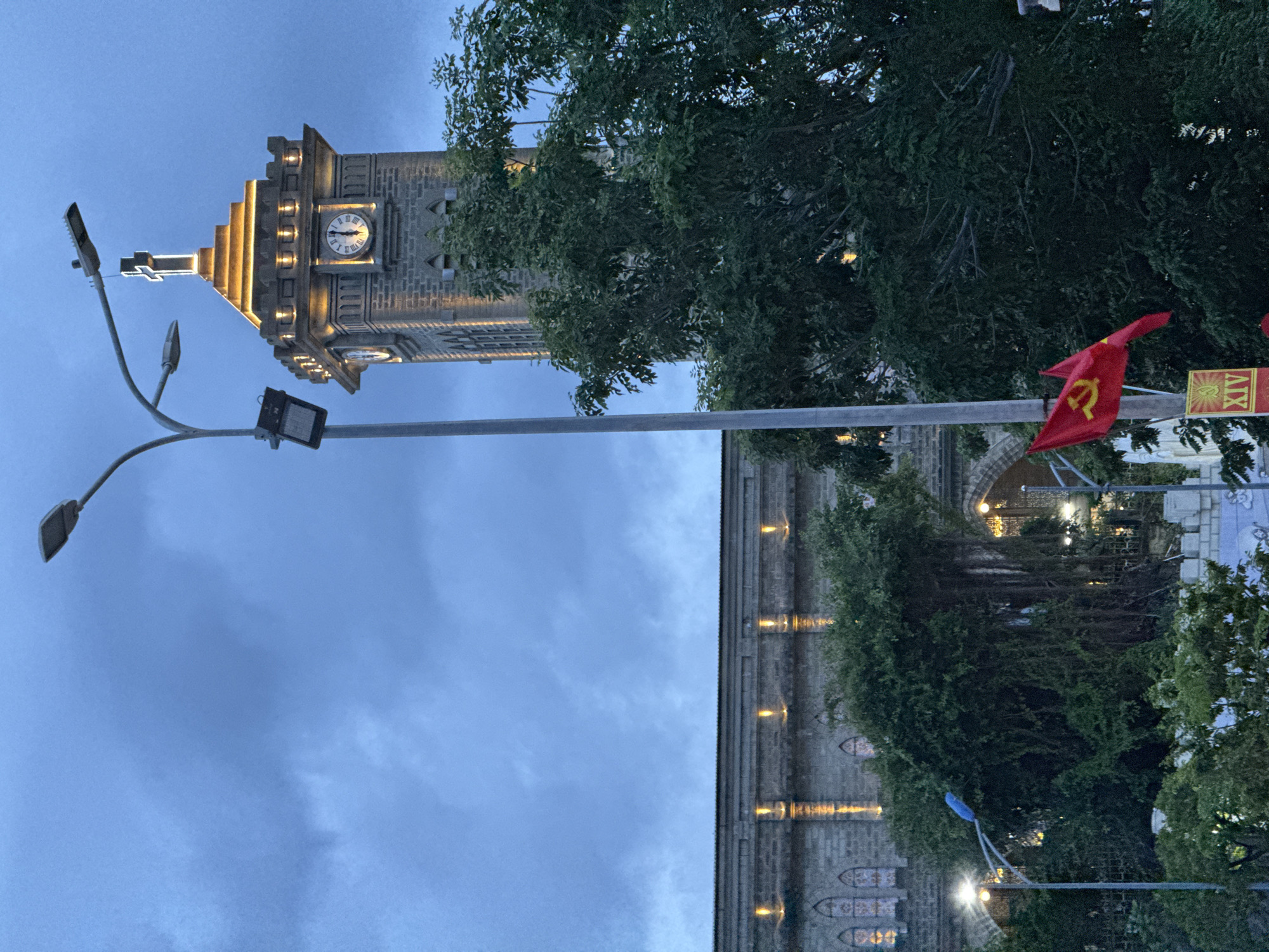 A church bell tower with a cross at the apex and illuminated by golden lights stands tall against a cloudy sky. The tower features a clock face with Roman numerals and two hands pointing to approximately 11:45. To the left of the tower, a long, grey lamppost with two lamps extends upwards, partially obscuring the tower. Adjacent to the lamppost is a stone building with illuminated arched windows. In the foreground, a large tree with dense green foliage partially obscures the lower part of the tower and the building. Two flags are visible: a red flag with a yellow hammer and sickle, and another flag with the same design on the left. A yellow sign with red Roman numerals "XIV" is also visible. The scene appears to be in Nha Trang, Vietnam, likely in the vicinity of the Nha Trang Cathedral, given the architecture of the building and the presence of the flags. The lighting suggests it is dusk or early evening.