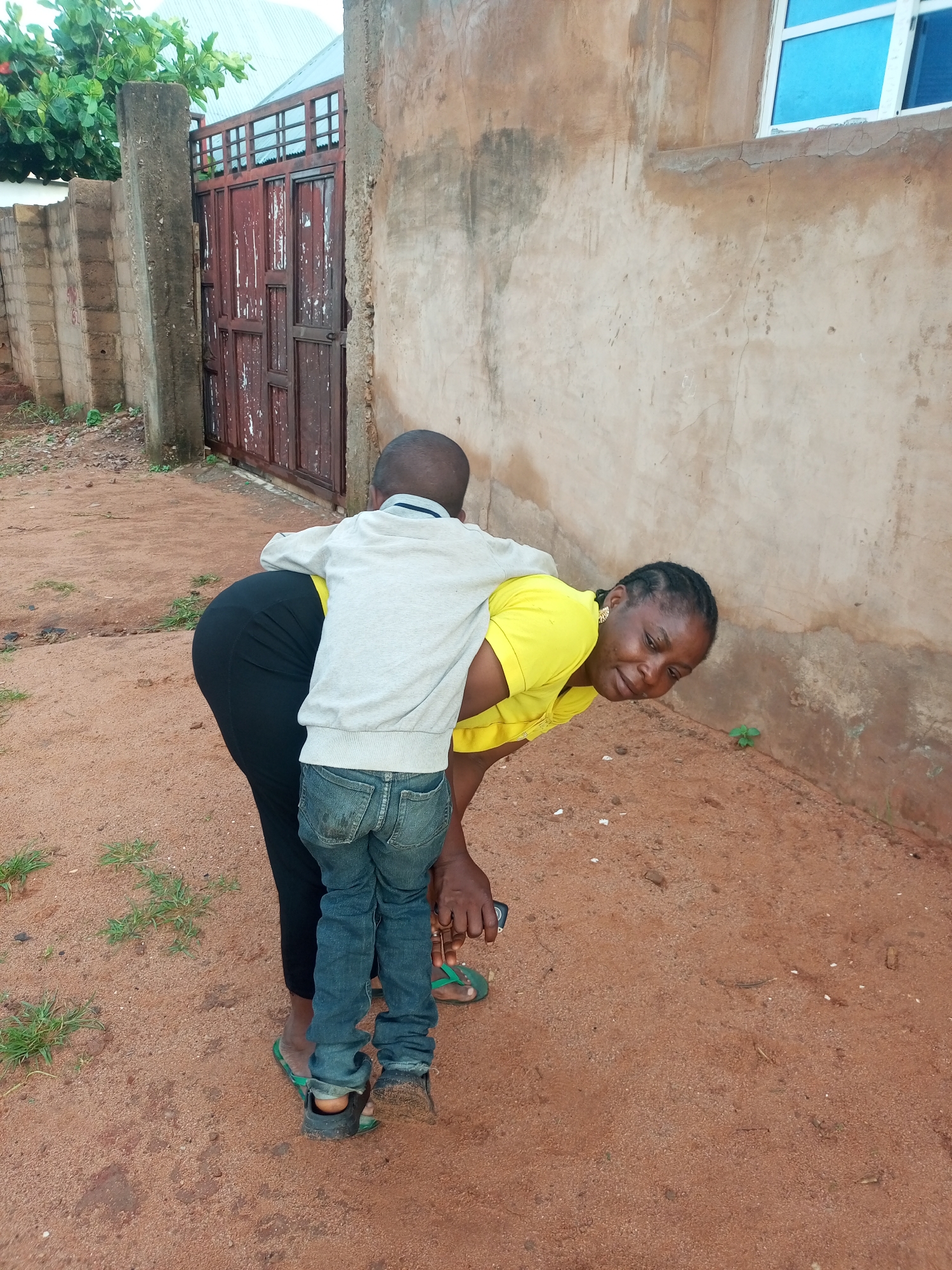 In Tempure, Nigeria, a woman in a bright yellow t-shirt bends down to assist a young boy wearing a light grey sweatshirt and blue jeans. The boy is embracing her from behind. They are standing on a sandy, reddish-brown ground with sparse patches of green grass.
Behind them, a weathered, dark red metal gate with a lattice design is visible, set against a rough, beige-colored wall of a building. To the left of the gate, a concrete pillar and a brick wall can be seen, with some green foliage above. To the right of the woman, the beige wall continues, showing signs of age with cracks and some darker discoloration. A portion of a building with a white-framed window featuring bright blue glass is also present on the right side of the frame.
The scene appears to be outdoors, possibly in a residential area, during daylight. The woman's expression suggests she is engaged in conversation or interaction with the boy, possibly with a hint of a smile or concern. The boy is holding onto her, indicating a close relationship or a moment of comfort. The overall atmosphere seems casual and domestic. No visible text is present in the image.